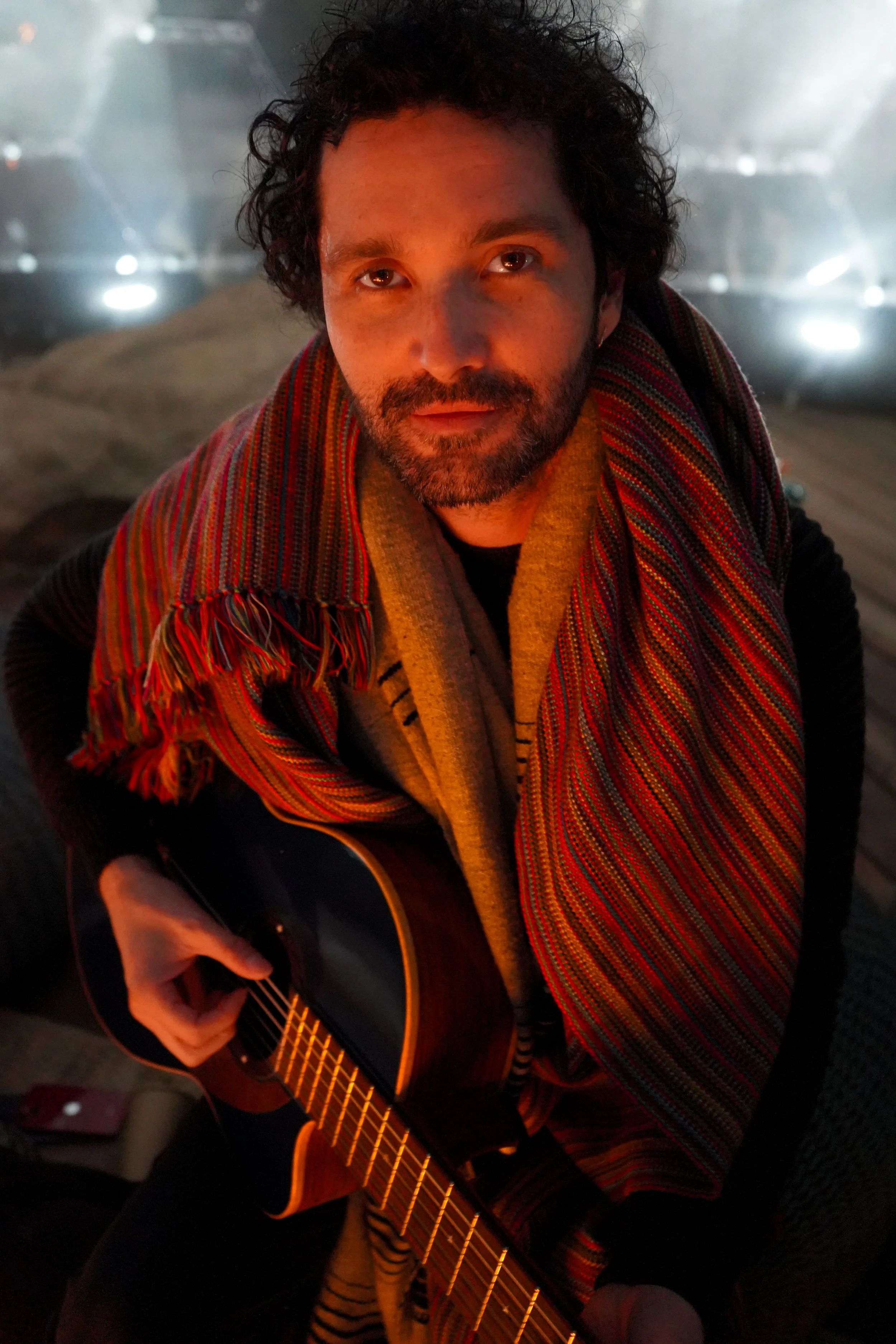 A man with curly hair and a beard, wearing a colorful striped scarf, is playing an acoustic guitar in a dimly lit room.