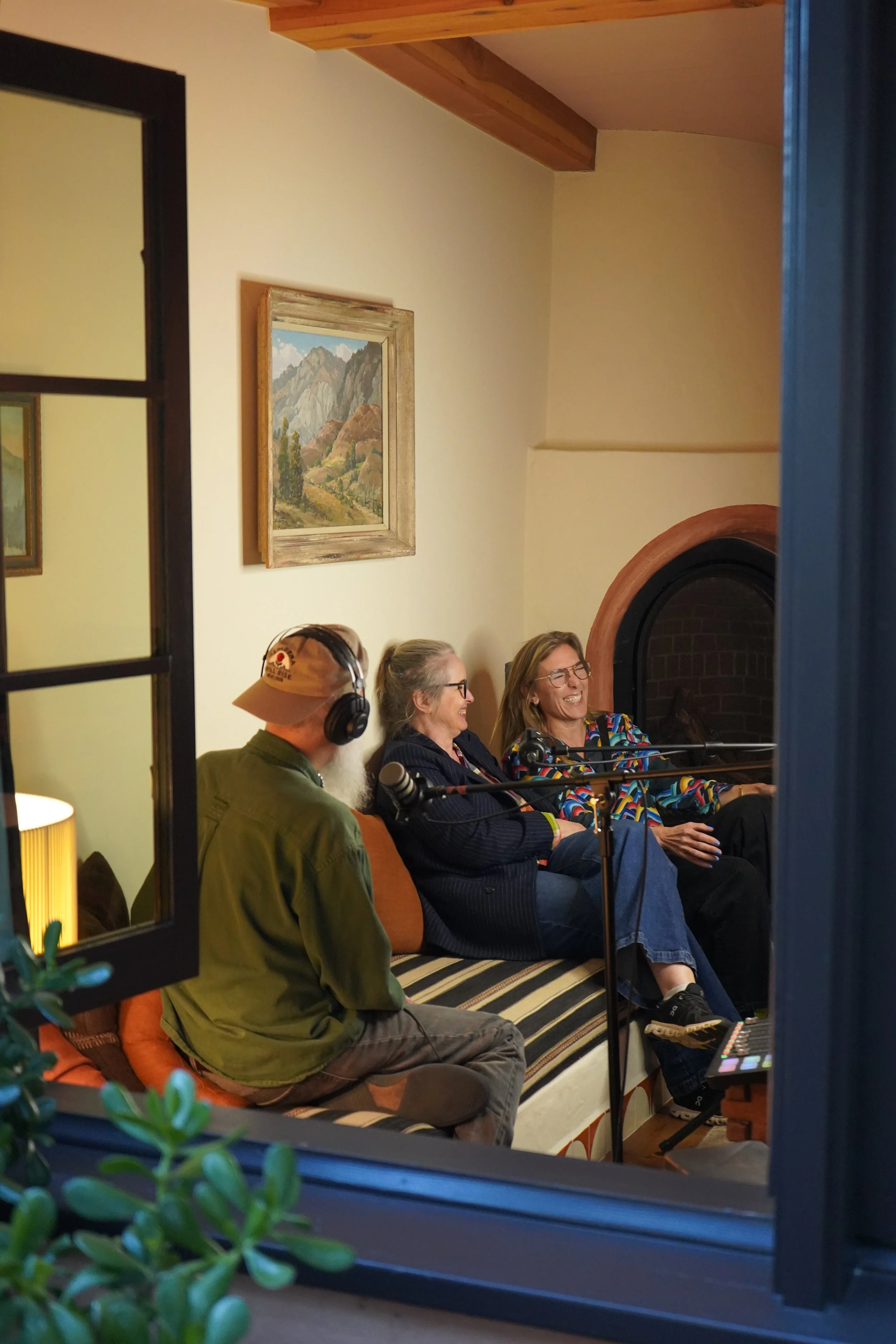 People participating in a podcast recording session in a cozy, warmly-lit living room with a fireplace, seen through a window.