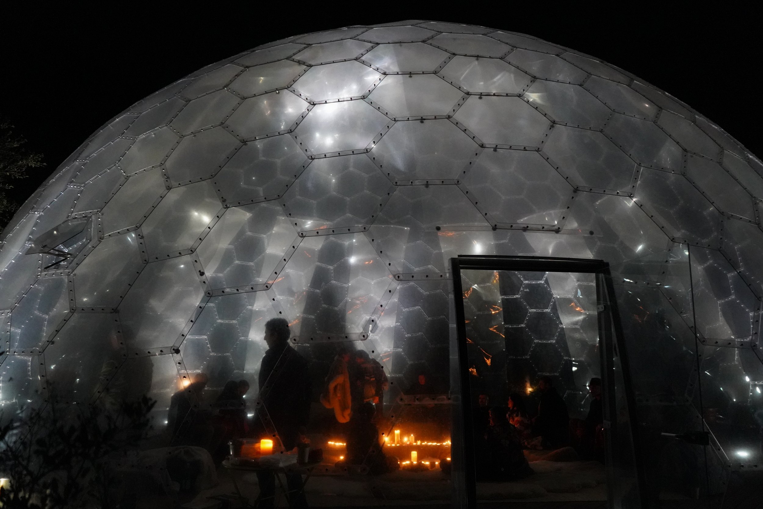 Nighttime scene of a geodesic dome with a transparent, hexagon pattern, illuminated from within, with silhouettes of people and candles inside.