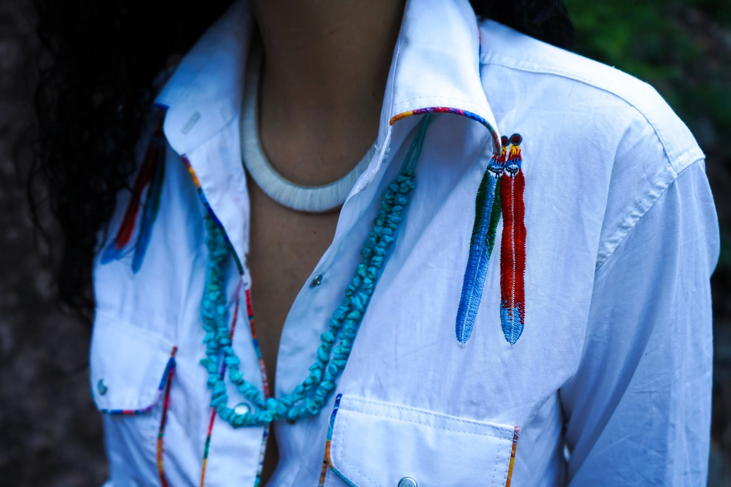 A person wearing a white button-up shirt with colorful embroidery of feathers and a beaded turquoise necklace.