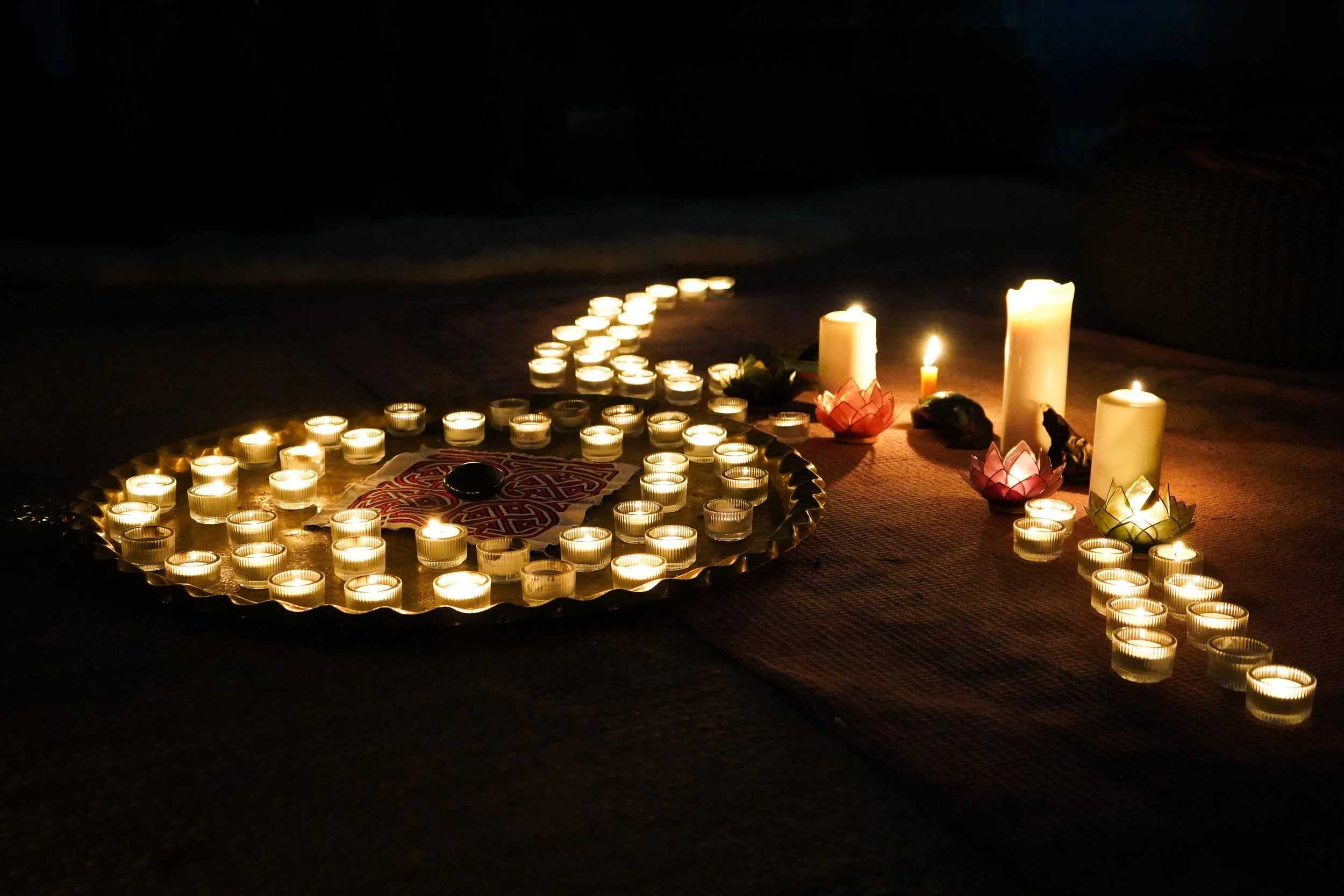 A display of lit candles, including tea lights and larger pillar candles, arranged on a dark surface with petals and decorative objects, creating a serene and peaceful ambiance.