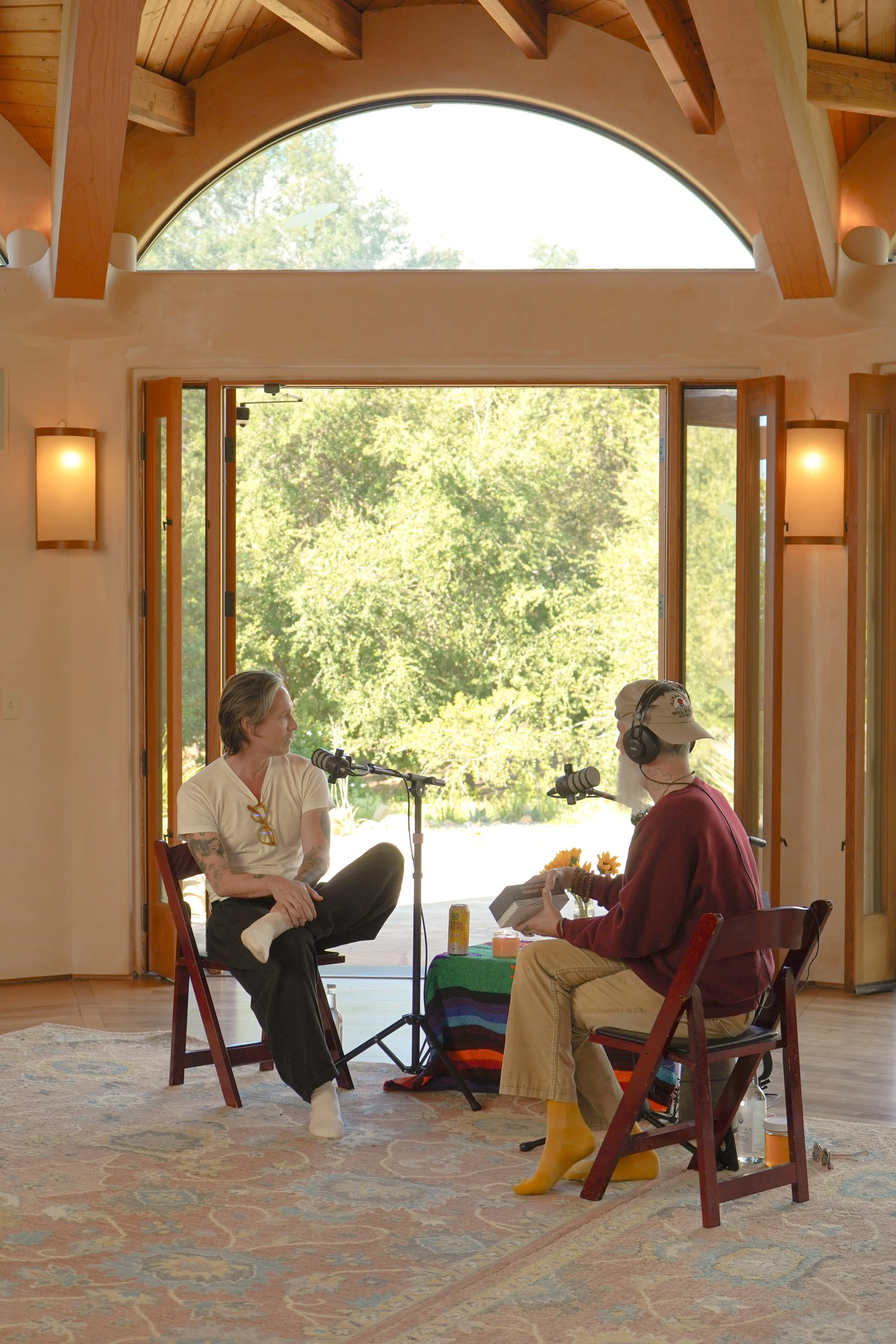 Two men having a podcast or radio interview inside a room with large glass doors opening to a scenic outdoor view of trees.