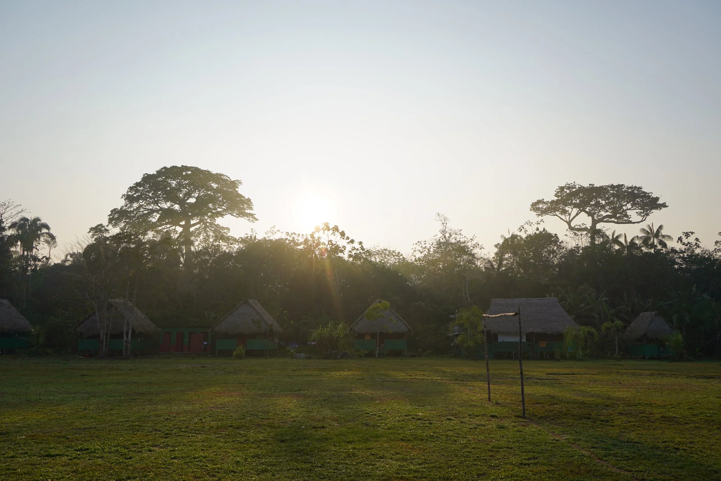 Sunset over small traditional huts with thatched roofs in a lush green landscape with tall trees in the background.