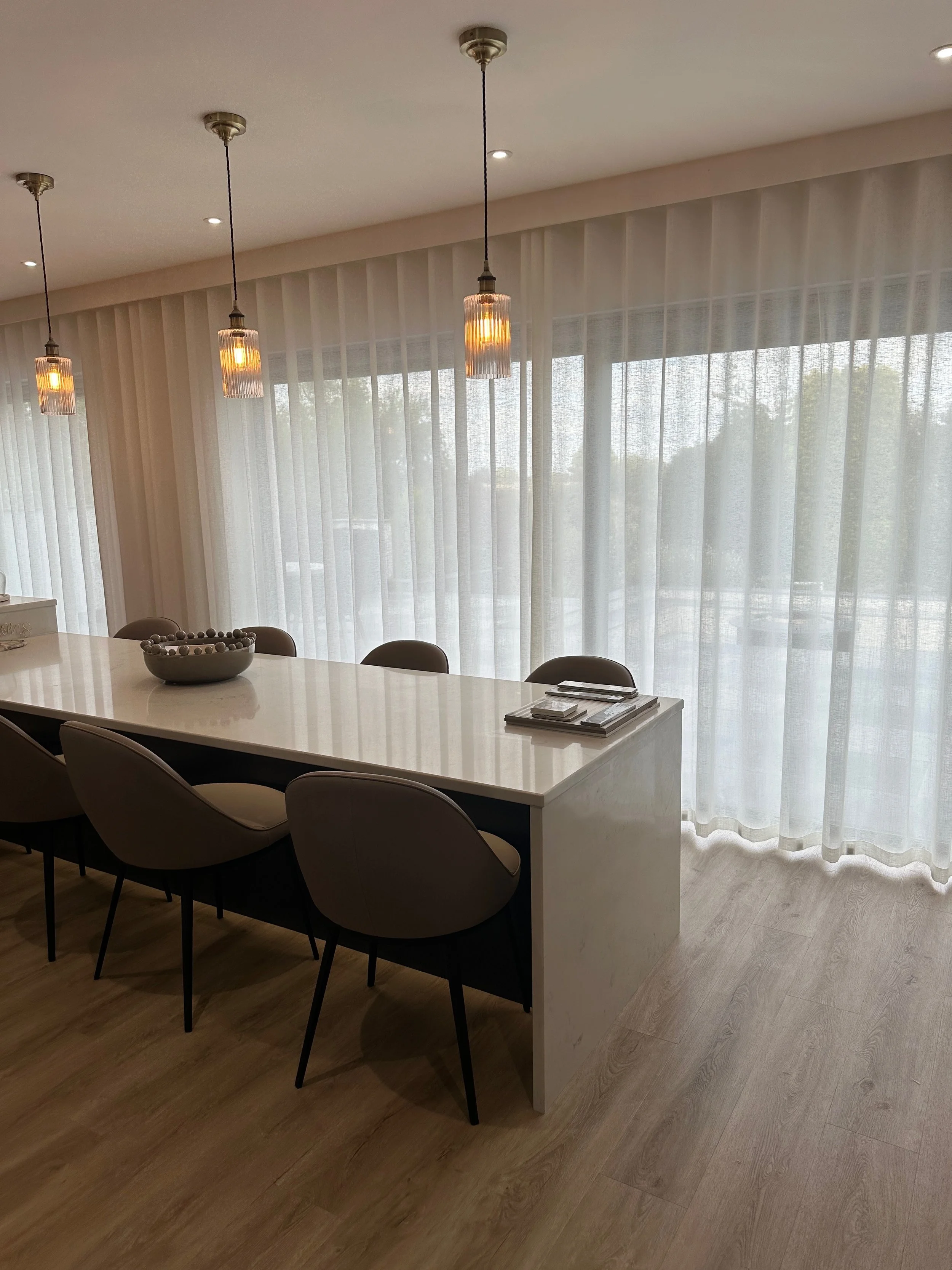 Modern dining room with a white marble table, six beige chairs, hanging pendant lights, sheer curtains, and wooden flooring.
