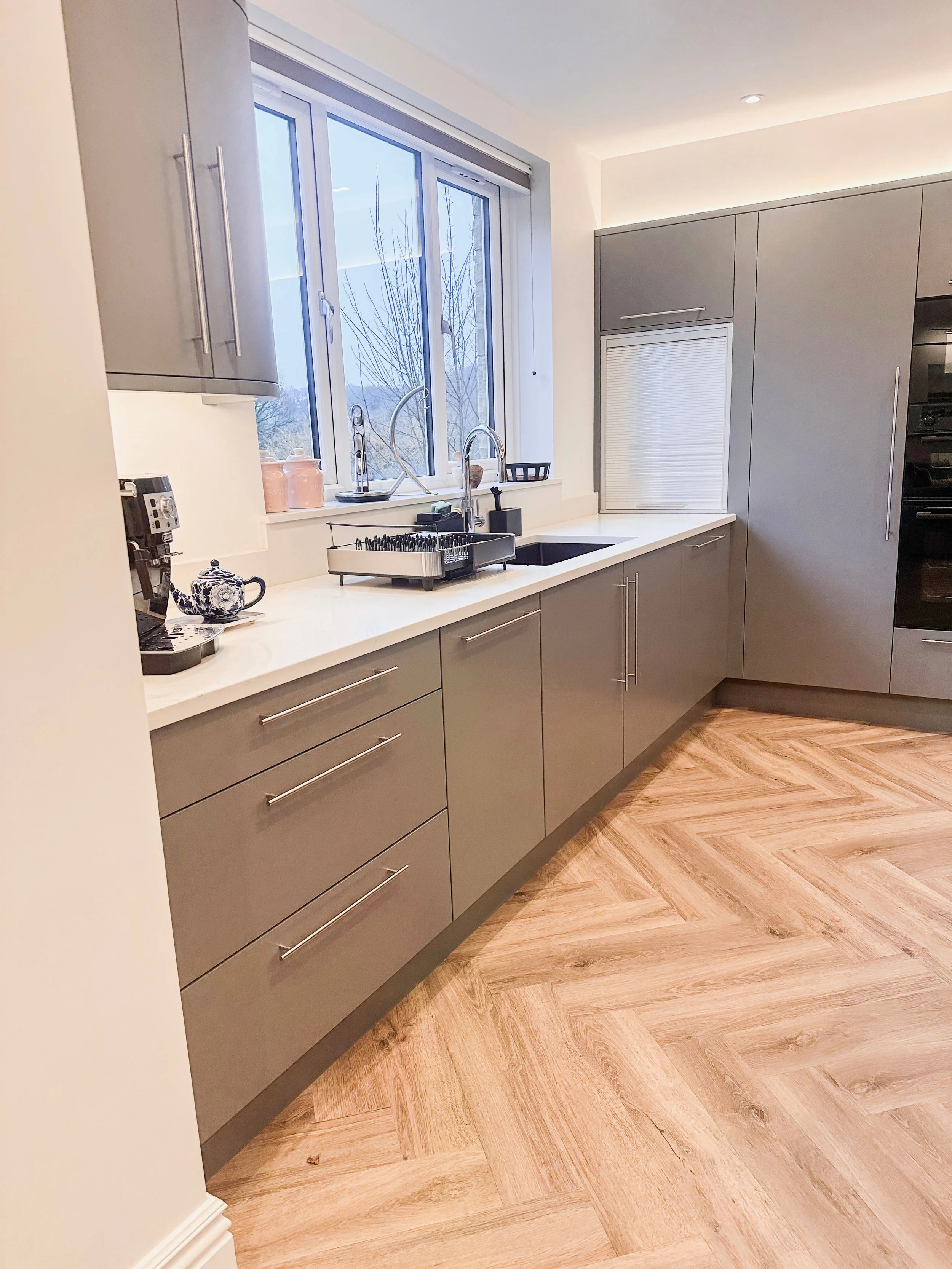 Modern kitchen with gray cabinets, white countertops, a window above the sink, and light wood flooring.