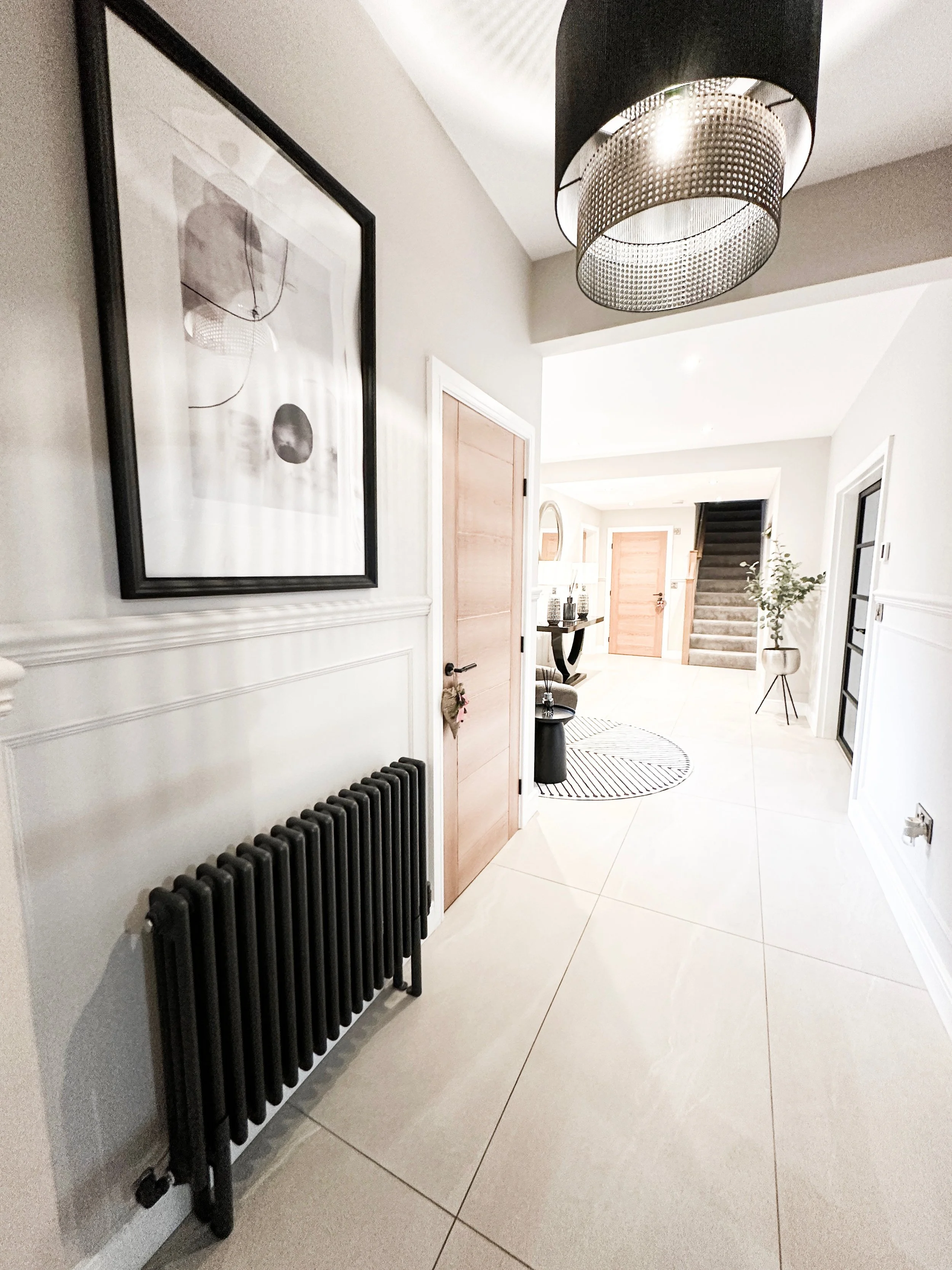 An entryway with light-colored tile flooring, a black radiator, a framed black-and-white art piece on the wall, a black and metallic hanging light fixture, a wooden door, a small black table with decorative items, a round striped rug, stairs with car