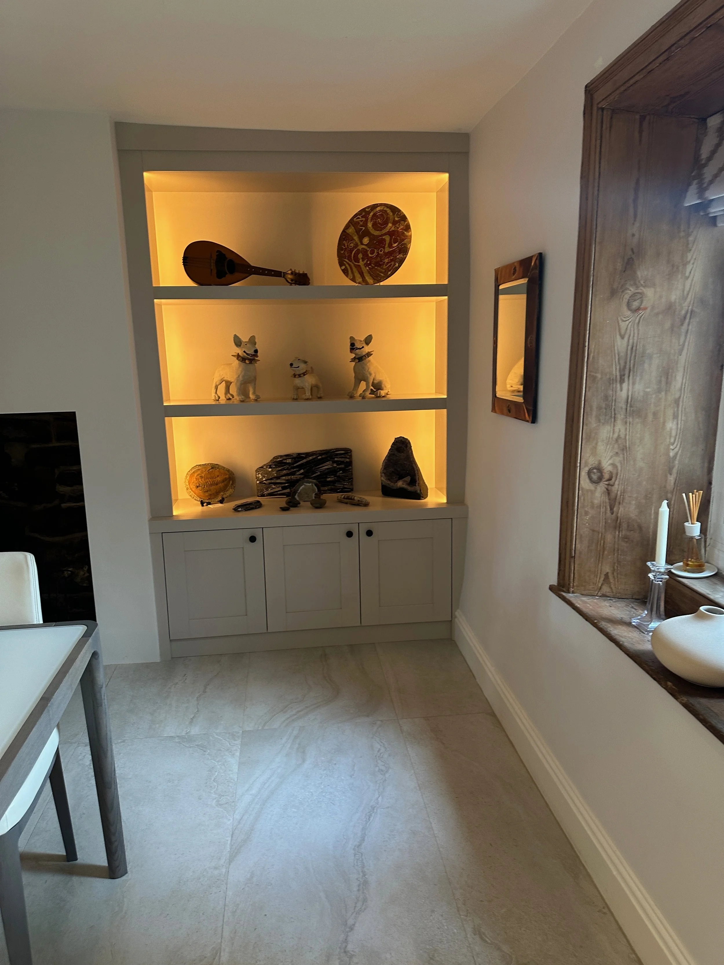Living room corner with built-in shelves illuminated with warm light, displaying decorative items including a guitar, pottery, dog figurines, and rocks. A wooden-framed mirror hangs on the wall, and part of a table with a white chair is visible.