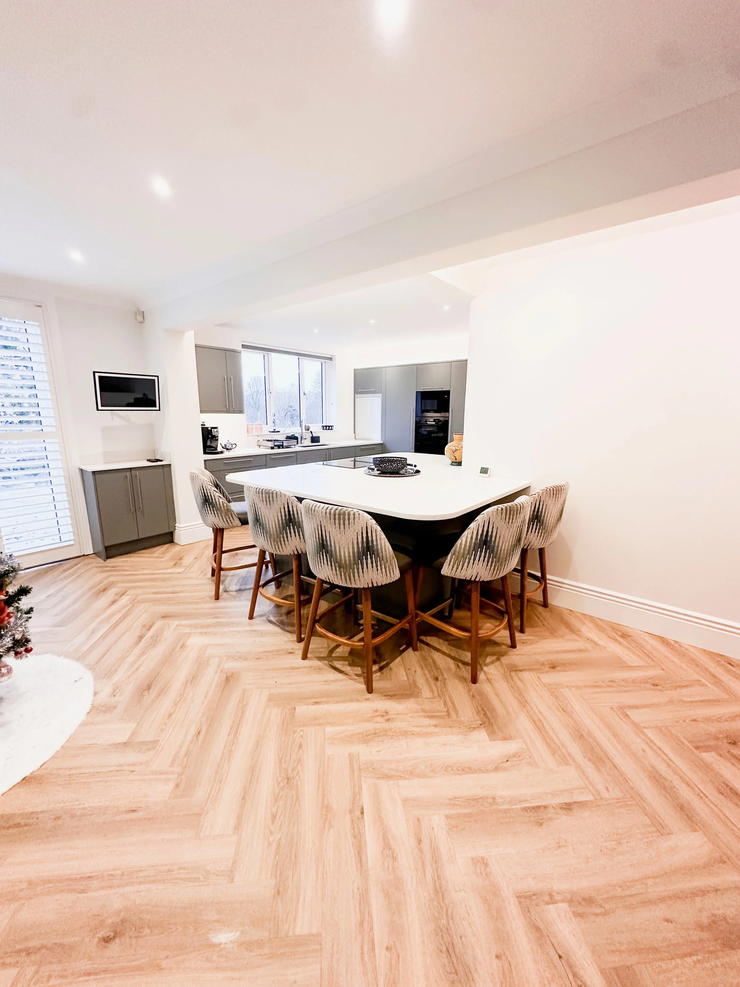 Bright, modern kitchen with a white island counter, gray cabinets, and wooden bar stools around the island. Light-colored wood herringbone flooring, windows with natural light, and a small wall-mounted TV.