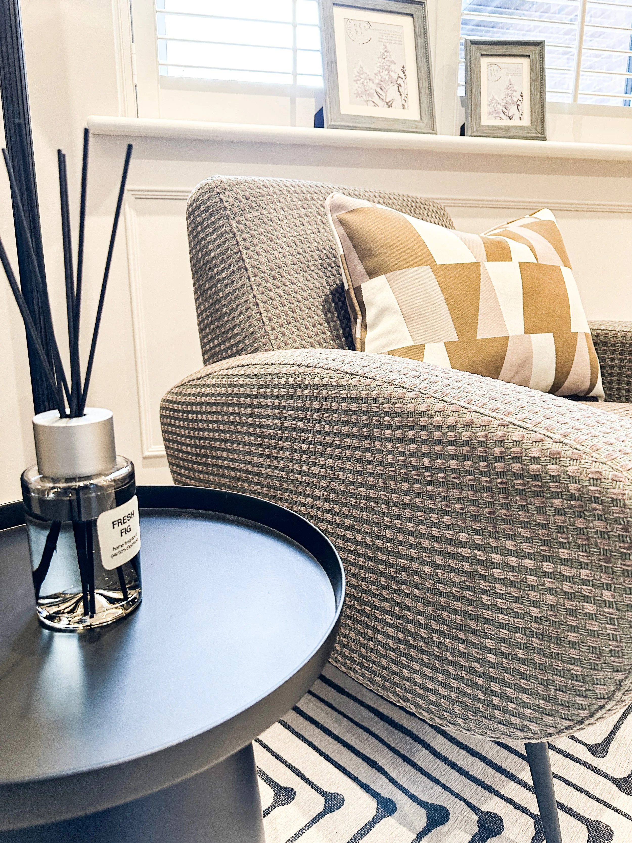 A living room corner with a textured armchair, a decorative pillow, a small black side table with a diffuser, and framed wall art above the window with white plantation shutters.
