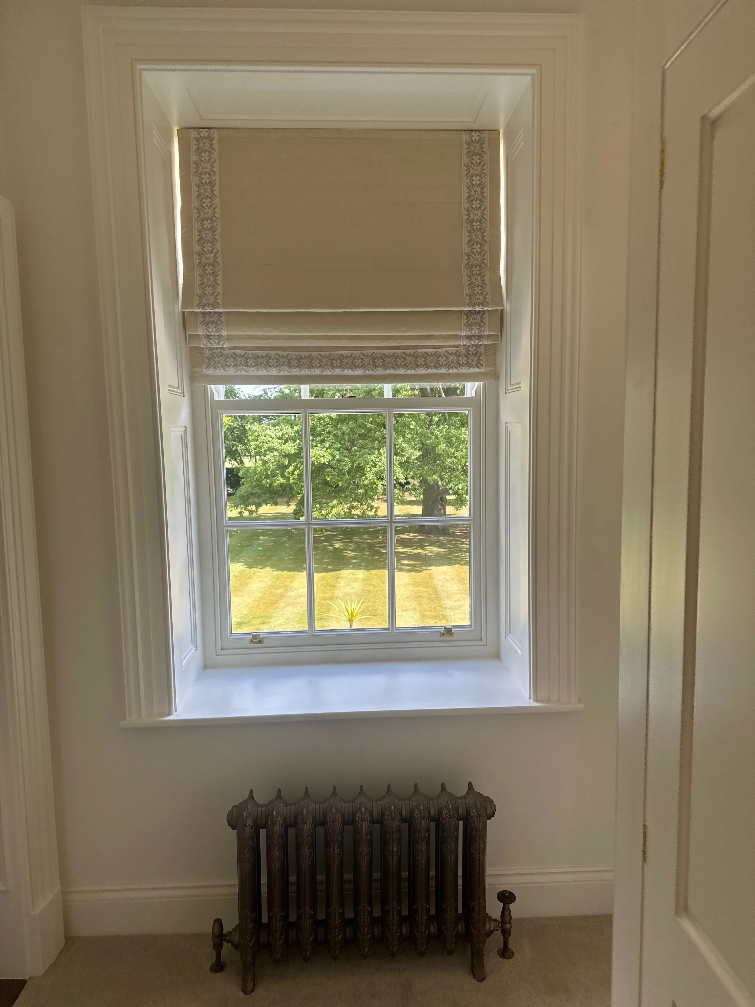 A window with valance blinds and view of green trees and grass outside, with an old radiator below.
