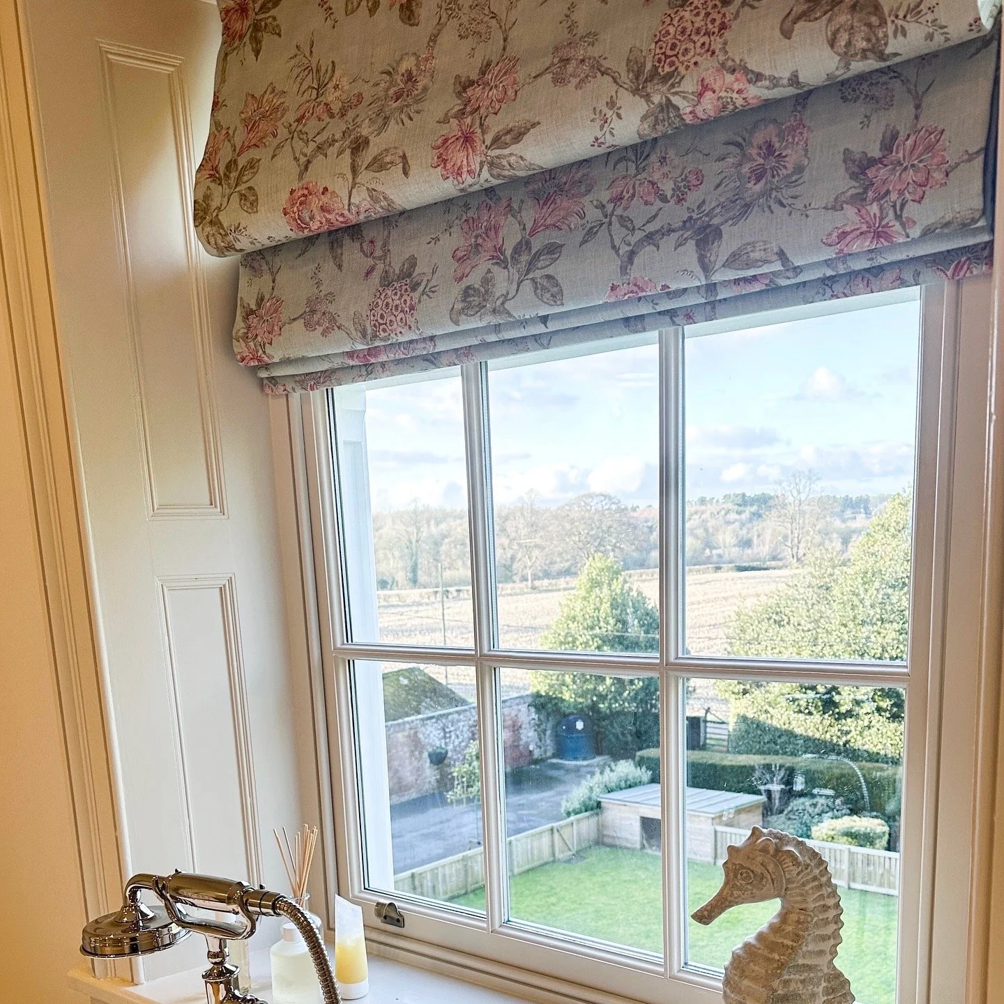 A kitchen window with floral valance curtains, offering a view of a backyard with grass, trees, a fence, and a cloudy sky. There is a ceramic seahorse sculpture and a bottle with reed diffusers on the windowsill, along with a small part of a silver f