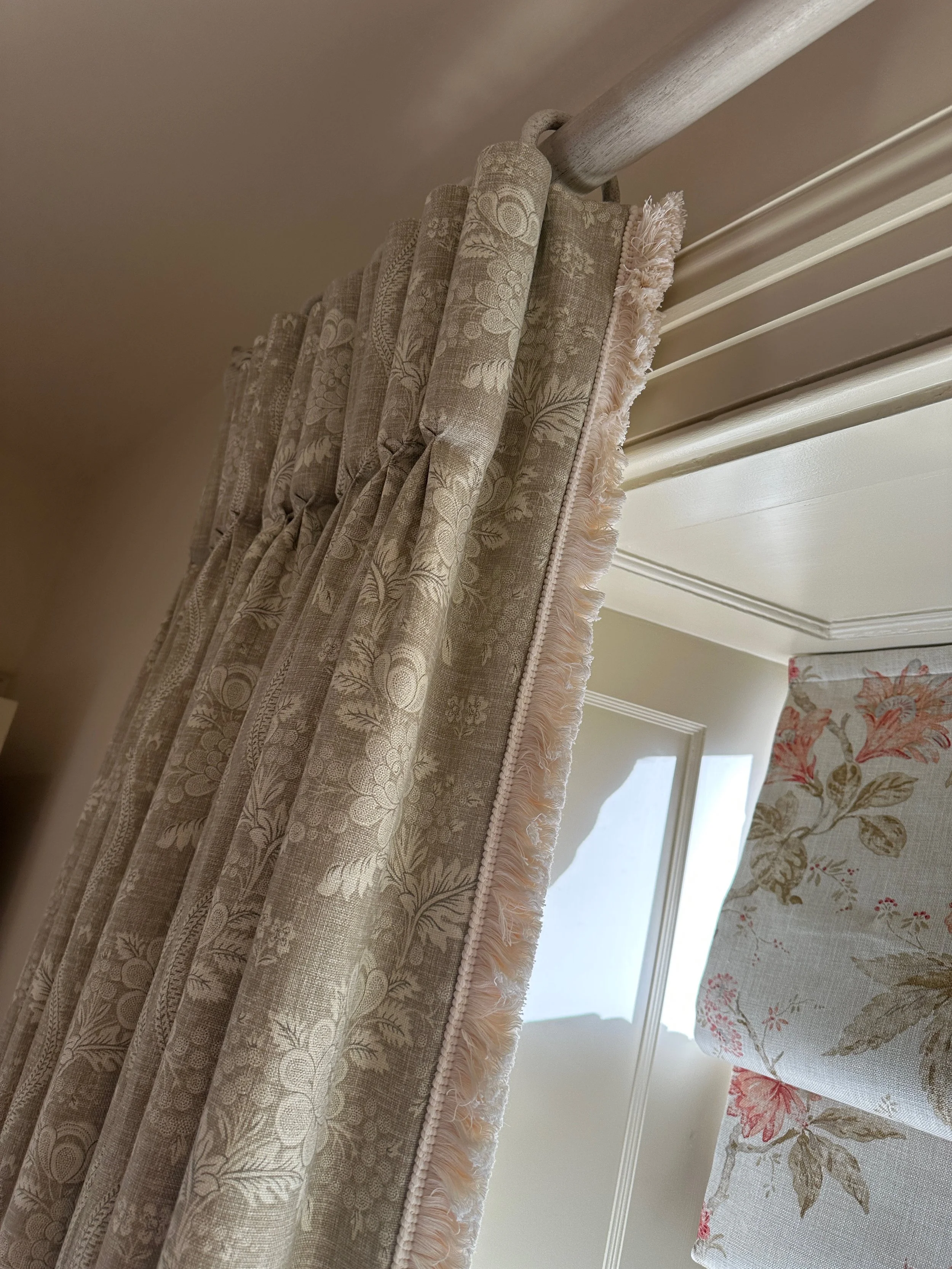 Close-up of patterned curtains hanging from a metal curtain rod near a window with white blinds and floral valance.