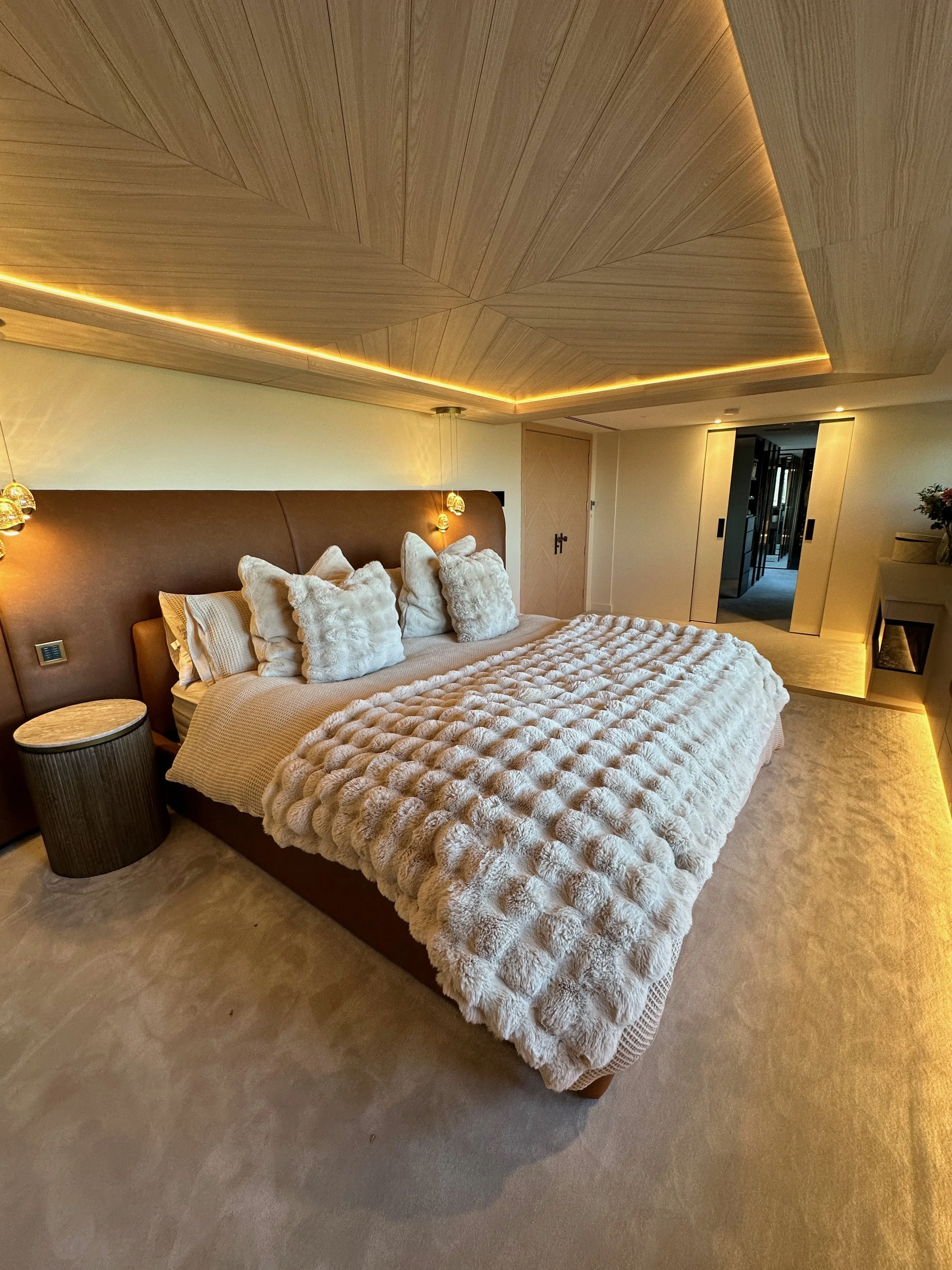 Elegant bedroom with a large bed, plush pillows, and a textured blanket, featuring wooden ceiling accents and ambient lighting.