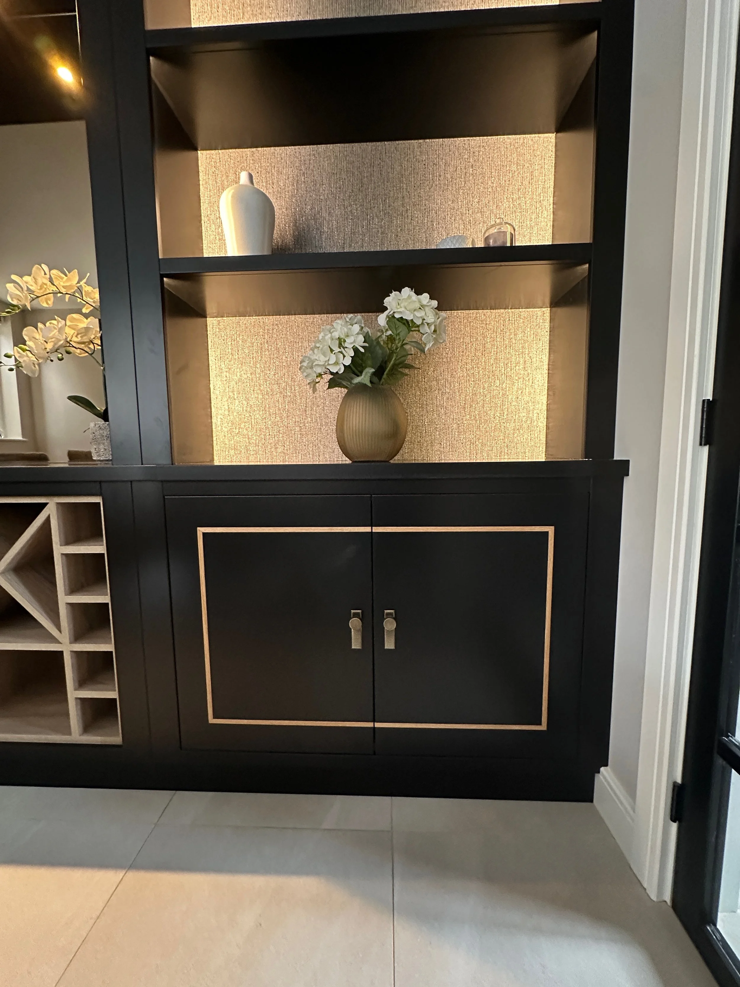 A black wooden cabinet with gold edging, featuring closed double doors, nestled in a corner next to a white wall door, with shelves above containing a beige vase with white flowers, and a bottle and small decor items on the top shelf. To the left, pa