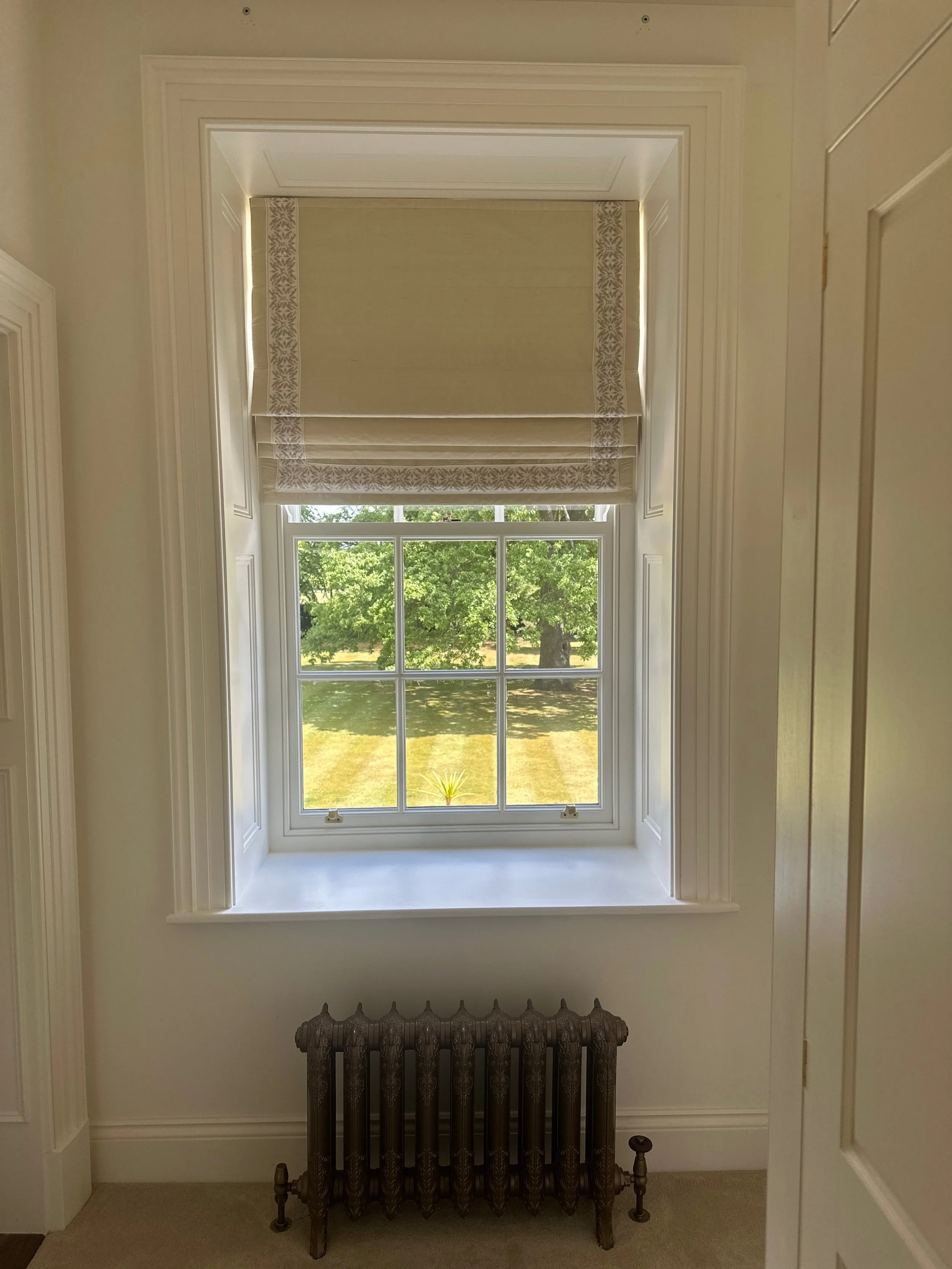 A window with a beige roman shade partially pulled up, revealing a view of green trees and grass outside. A vintage cast iron radiator is beneath the window, and the interior walls are painted light cream with white trim.