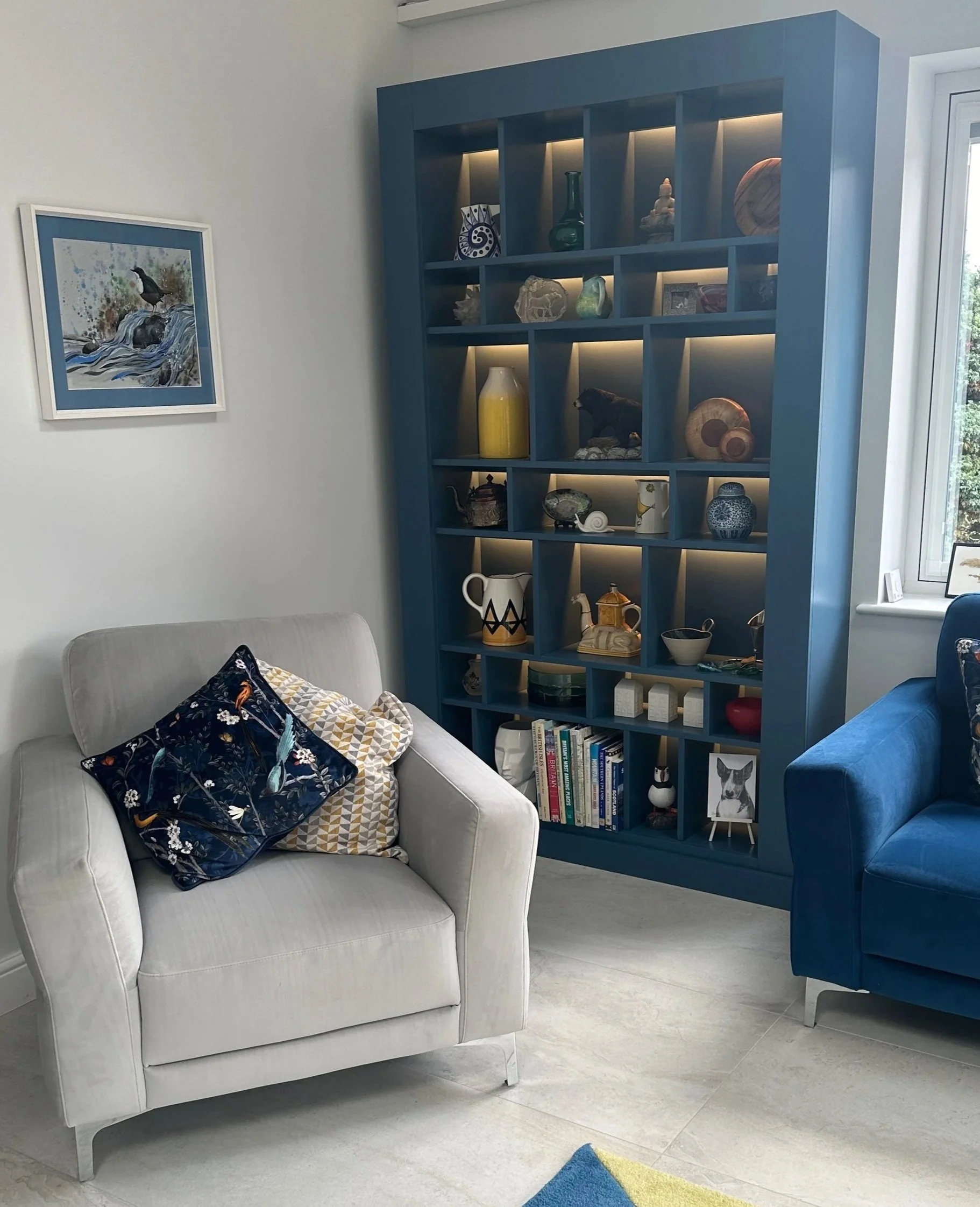 Living room corner with a white armchair with pillows, a blue armchair, and a large blue display shelf filled with decorative objects, books, and art pieces near a window.