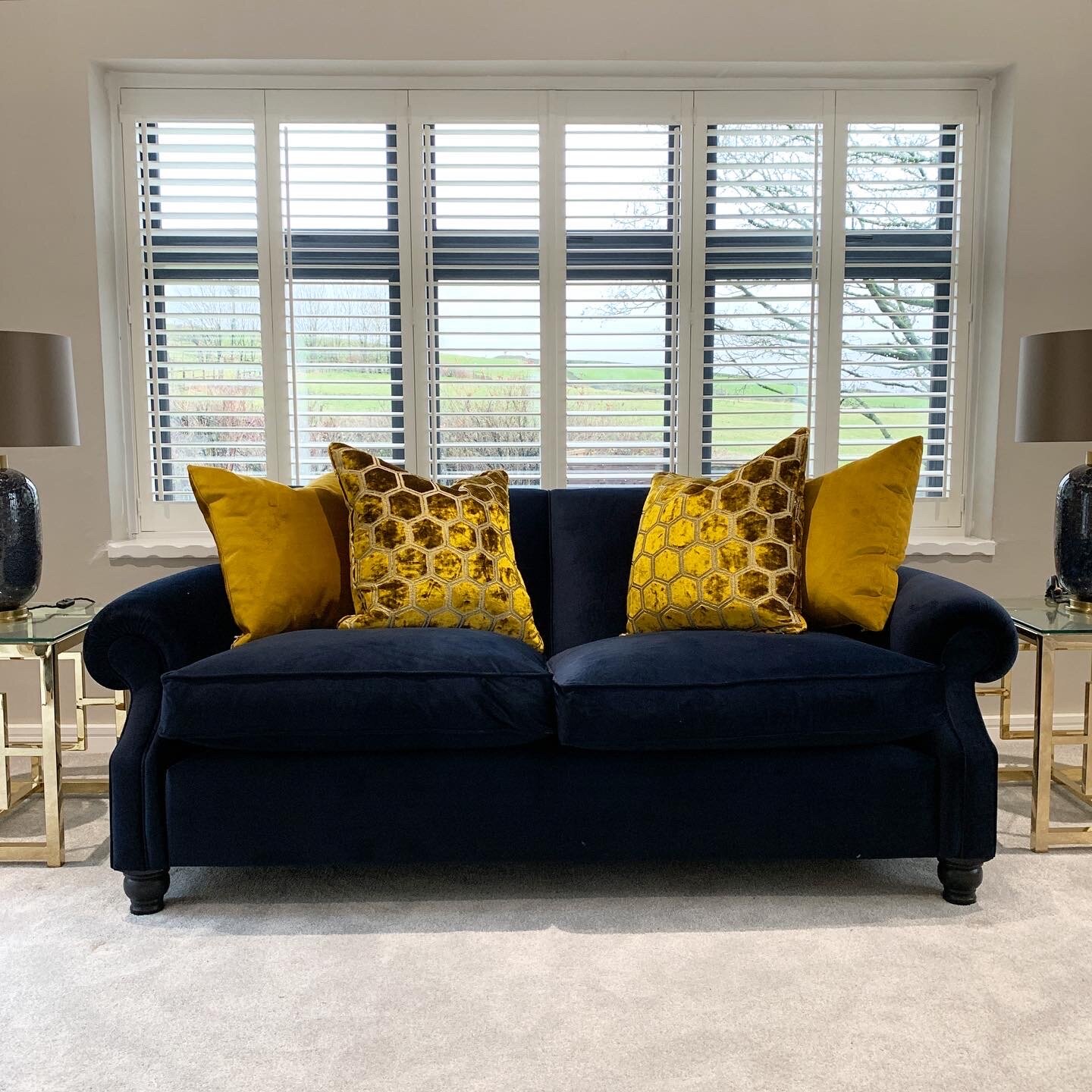 A dark blue velvet sofa with four yellow and gold pillows, placed in front of a large window with white blinds, flanked by two glass side tables with black lamps, in a living room.