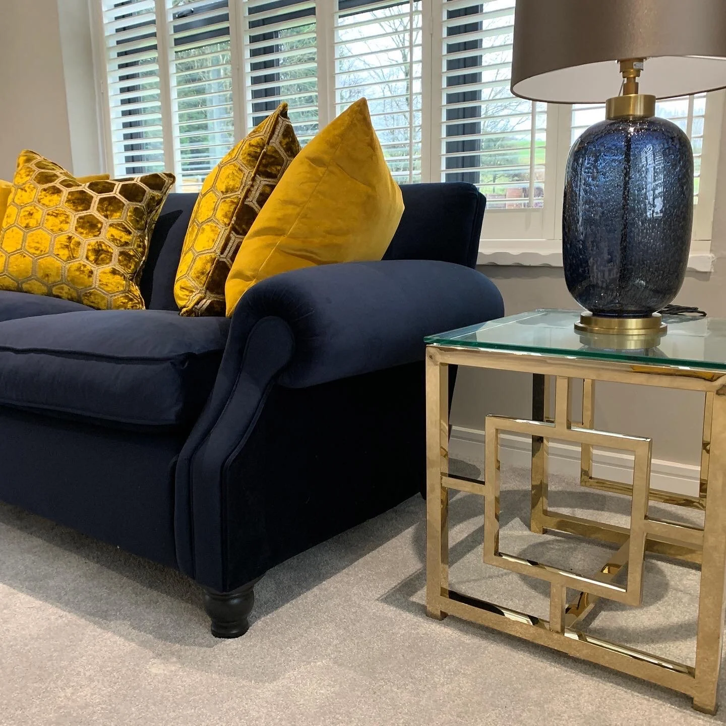 A dark blue sofa with three yellow throw pillows, a gold framed glass side table, and a blue ceramic table lamp with a white shade, in front of a window with white blinds, and a view of trees outside.