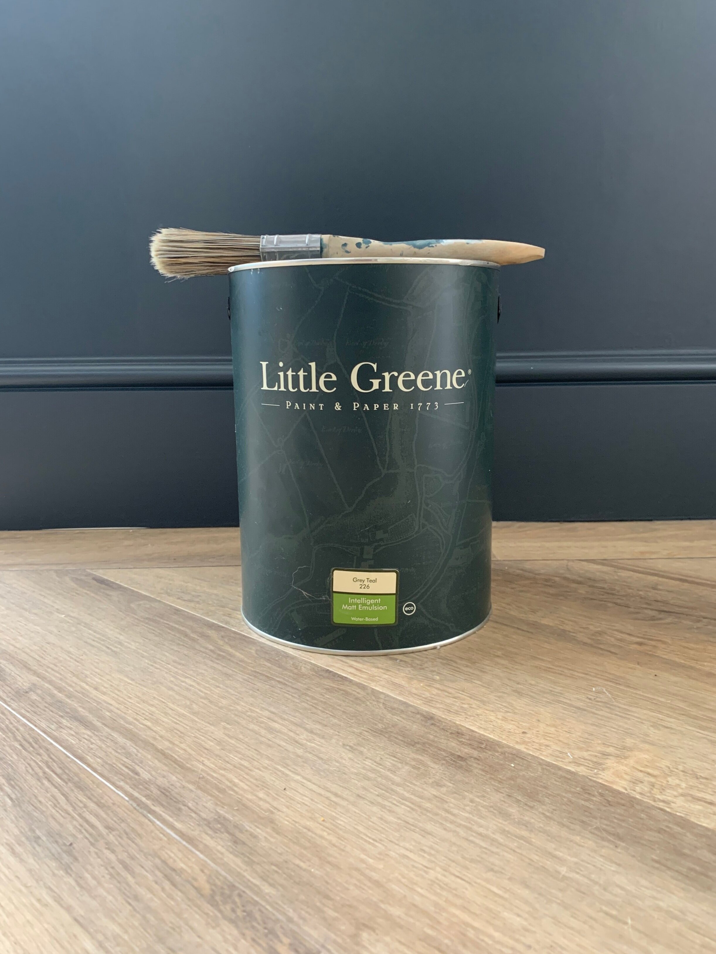 A can of Little Greene paint with a paintbrush resting on top of it on a wooden surface.