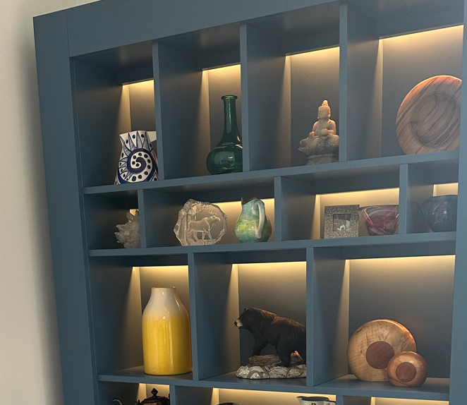 A blue shelving unit with multiple compartments, illuminated by warm lighting, displaying decorative objects including sculptures, vases, and artwork.