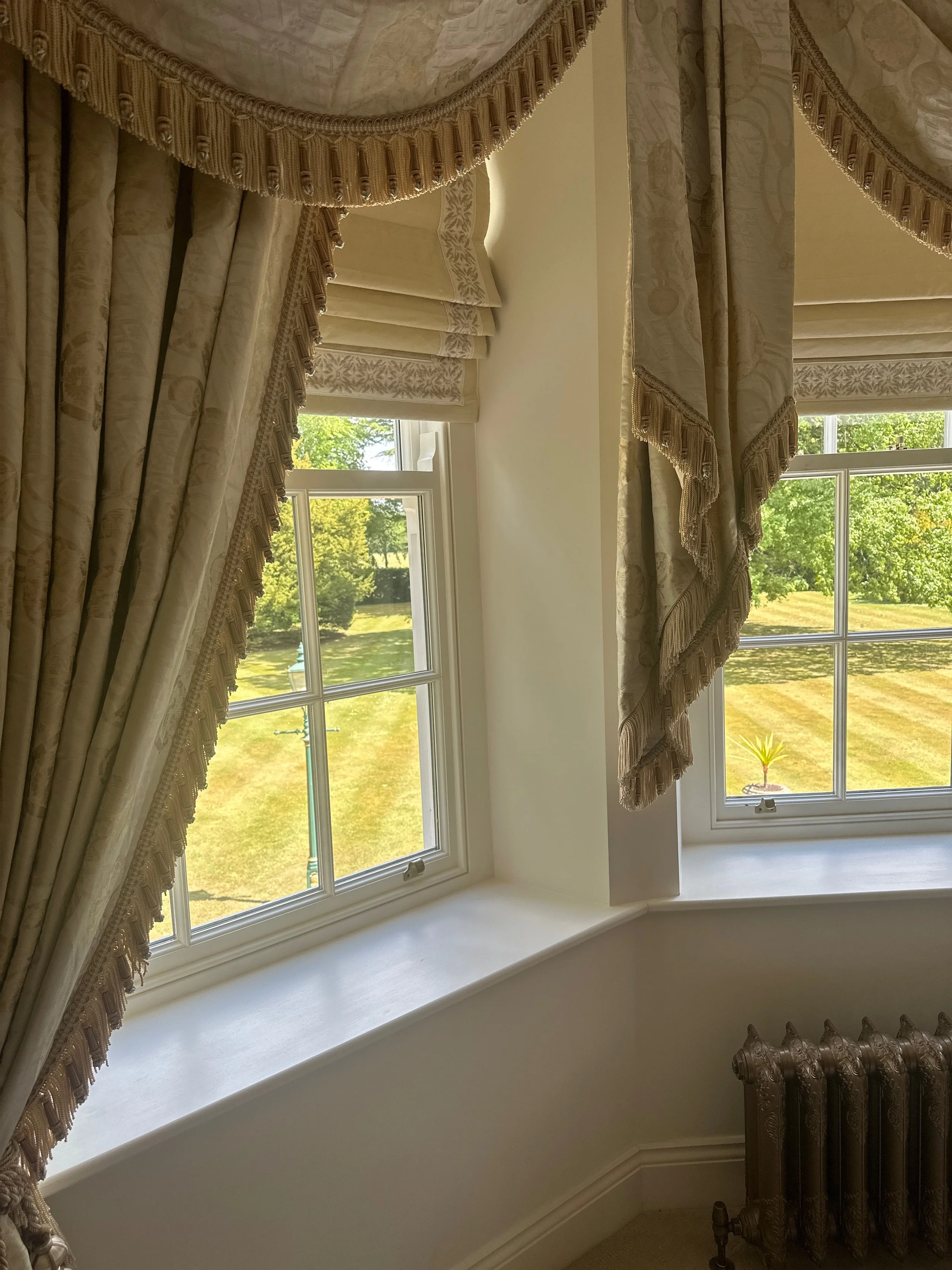 Interior view of a room showing decorative curtains with tassels hanging over two windows, with a view of a grassy yard and trees outside.