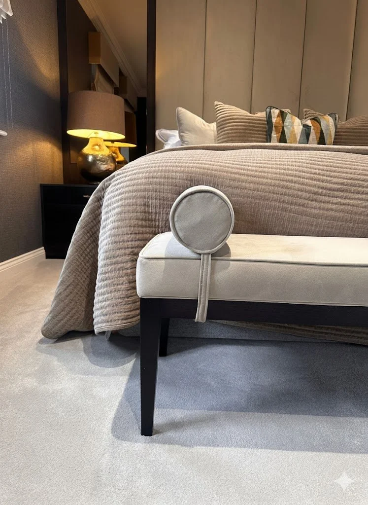 A bedroom with a bed, decorative pillows, a brown textured bedspread, bedside table, and lamp. A small round gray purse hangs on the bed's corner.