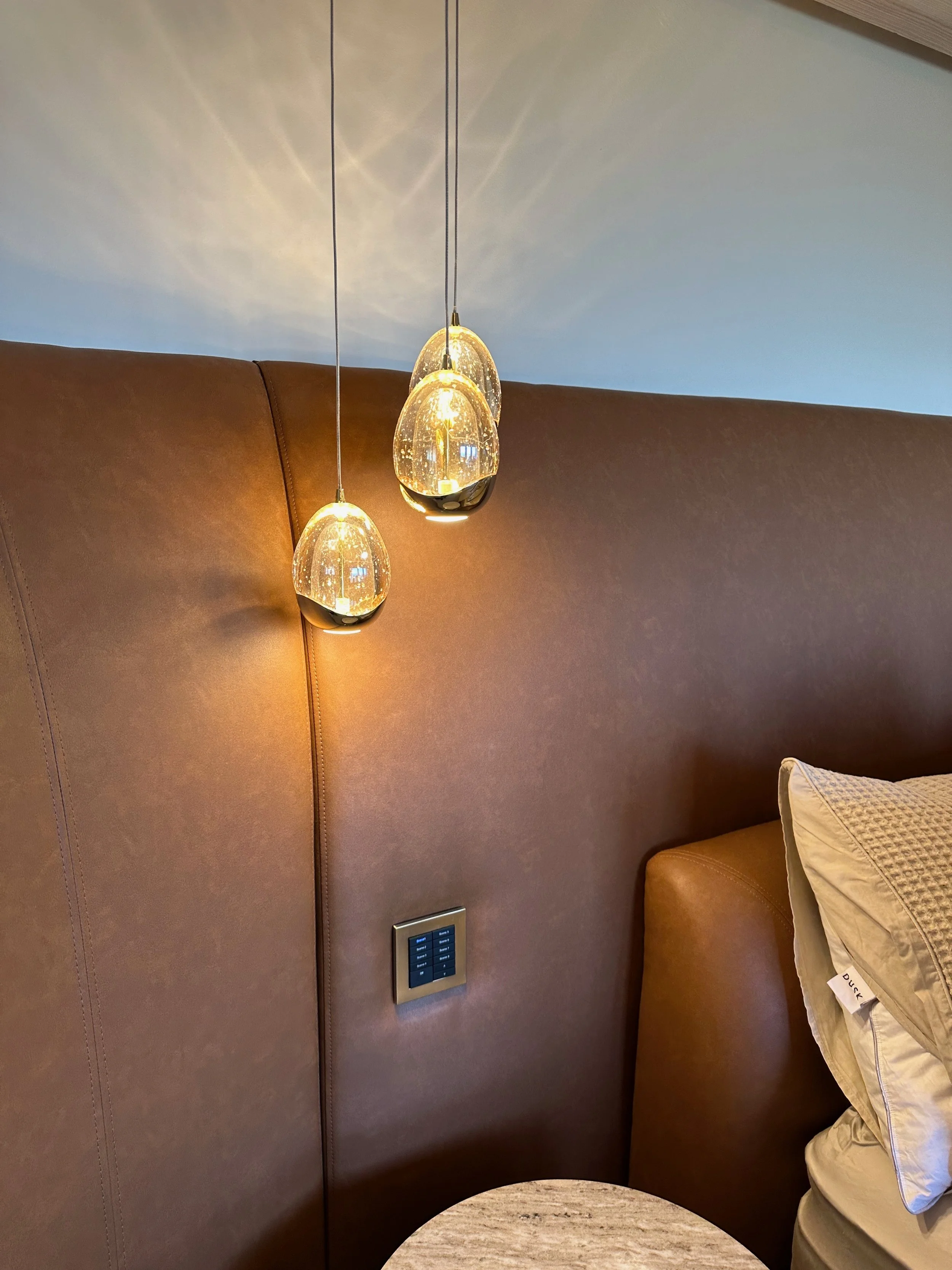 Modern bedroom with a large brown upholstered headboard, three pendant lights hanging from the ceiling, and a pillow on the bed.