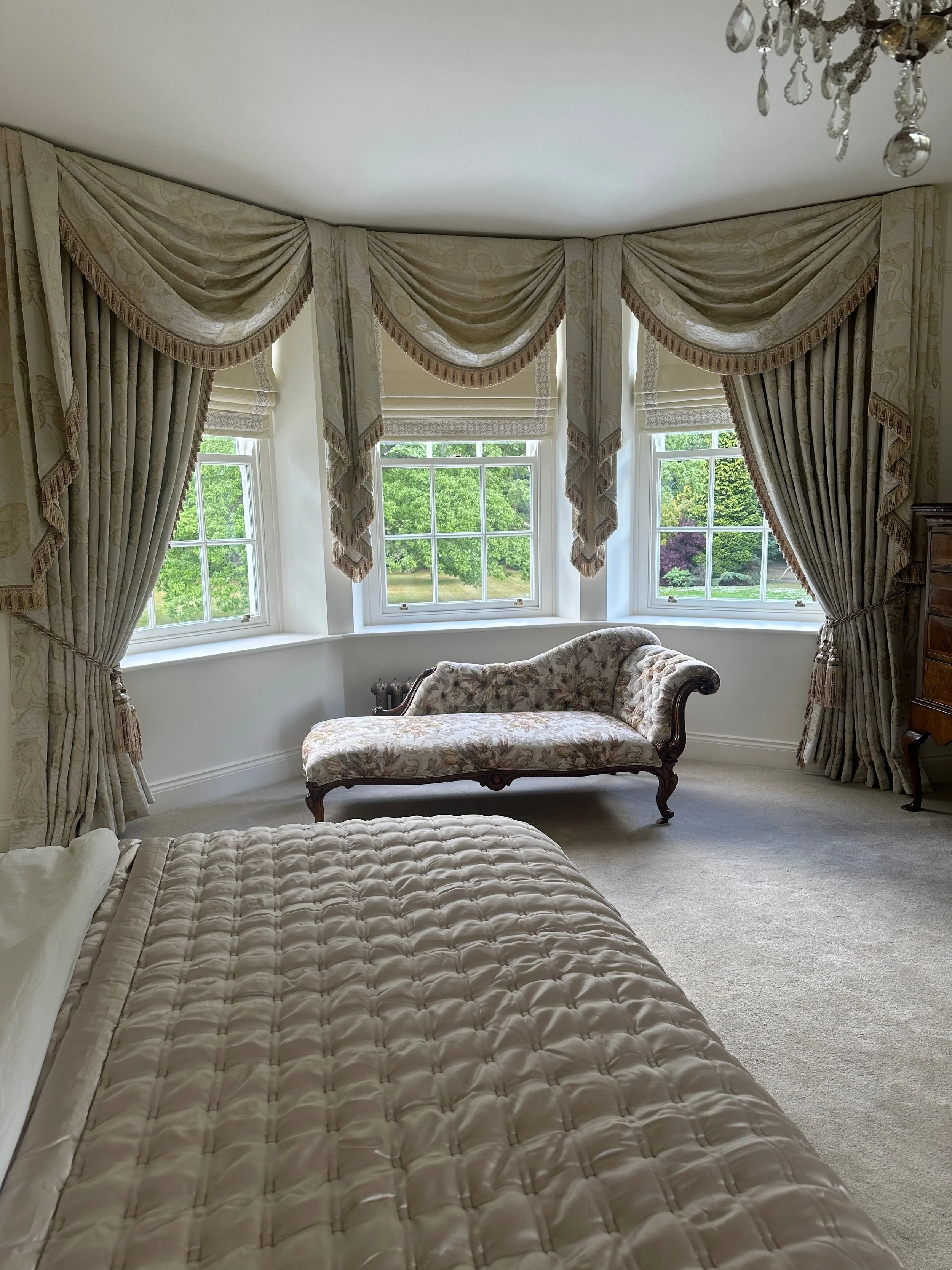 Bedroom with large windows, ornate curtains, and a floral upholstered chaise lounge.