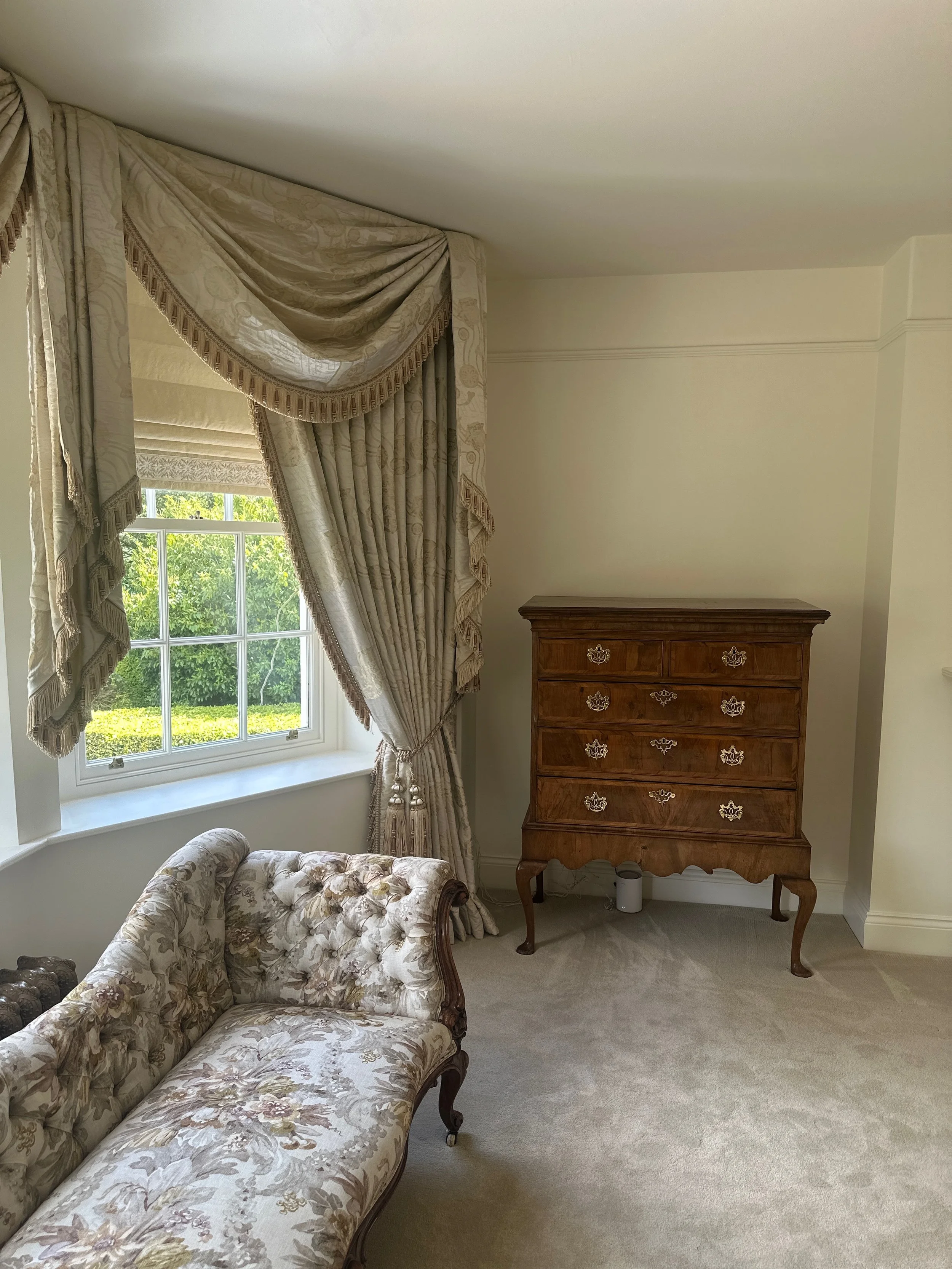 Living room with cream-colored walls, ornate floral upholstered sofa, window with elegant drapes, and a wooden chest of drawers.