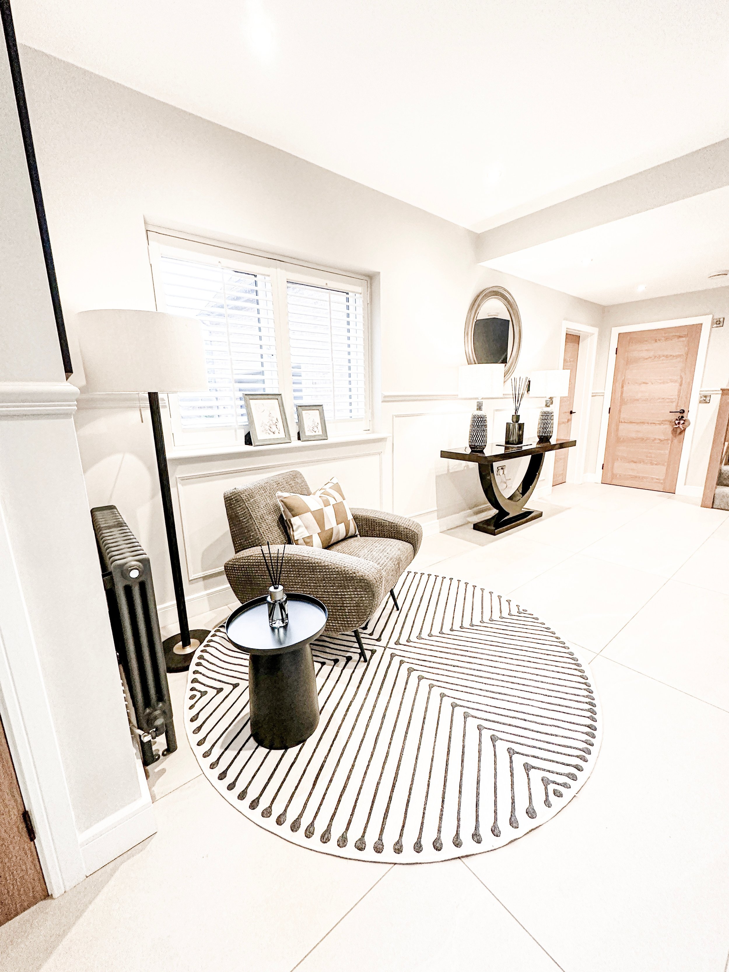 A modern, minimalistic living room corner with a round black and white striped rug, a small brown armchair, a black side table with reed diffuser, white window blinds, a tall floor lamp, decorative framed pictures, and a black console table with lamp