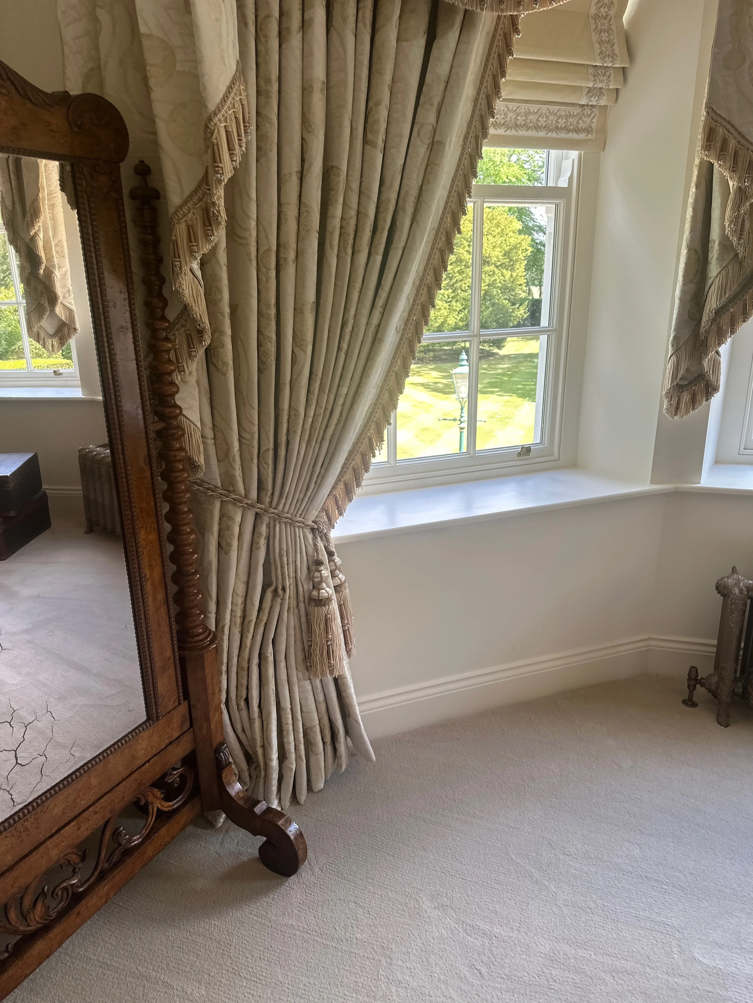 Elegant bedroom with large window, ornate curtains, and a wooden mirror frame.
