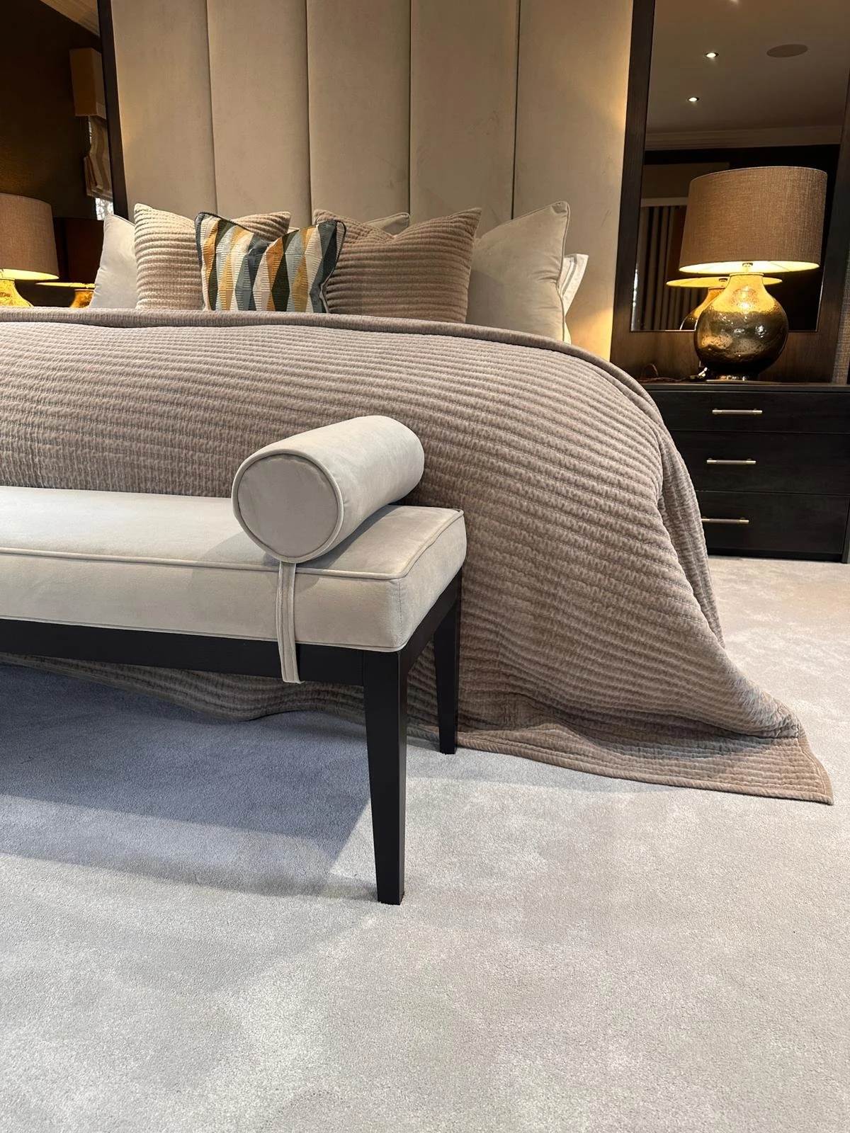 A bedroom with a bed covered in beige textured bedding and multiple pillows, a bench at the foot of the bed, and a nightstand with a lamp.