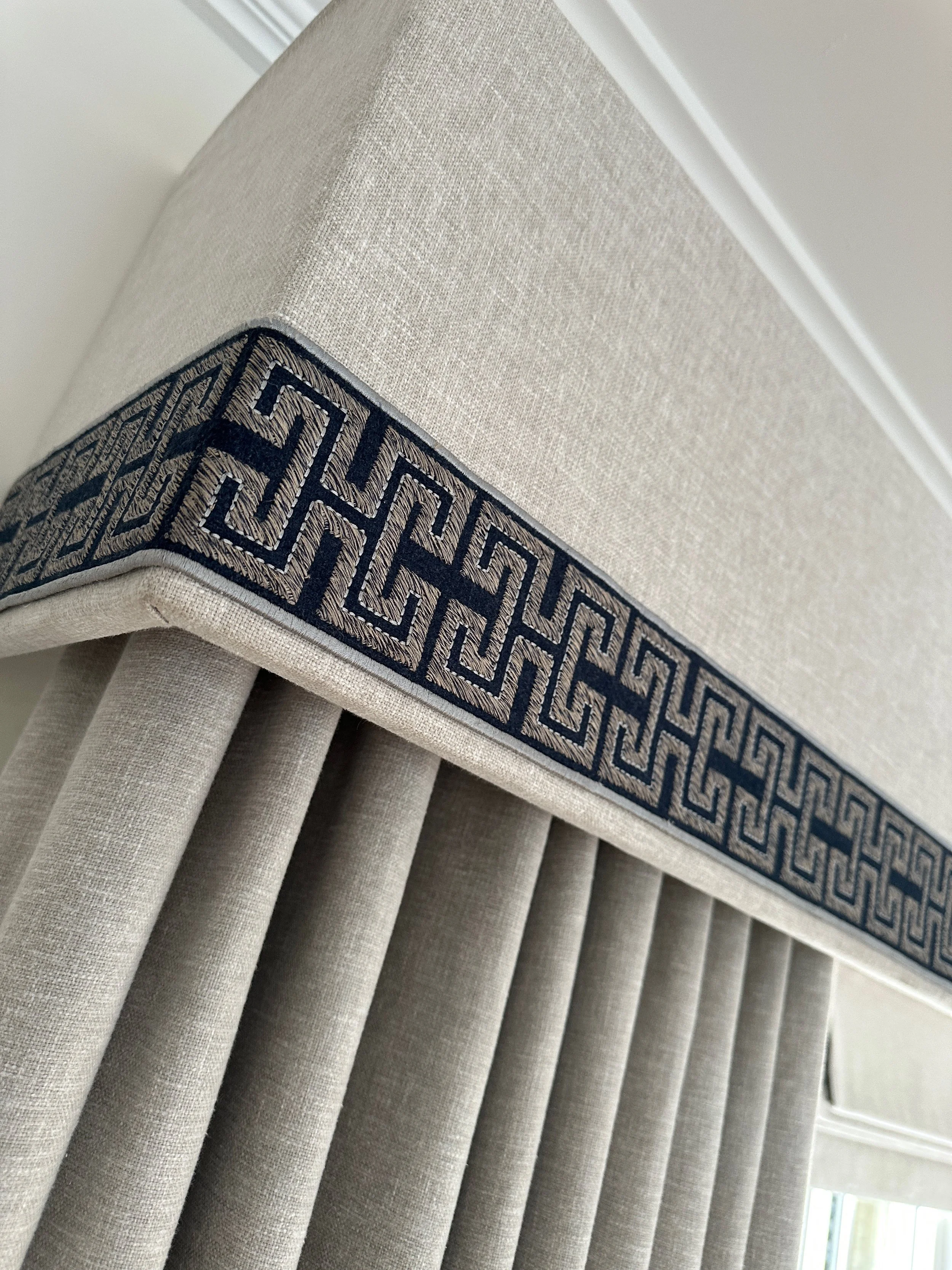 Close-up of a beige upholstered window valance with a dark embroidered Greek key pattern trim.