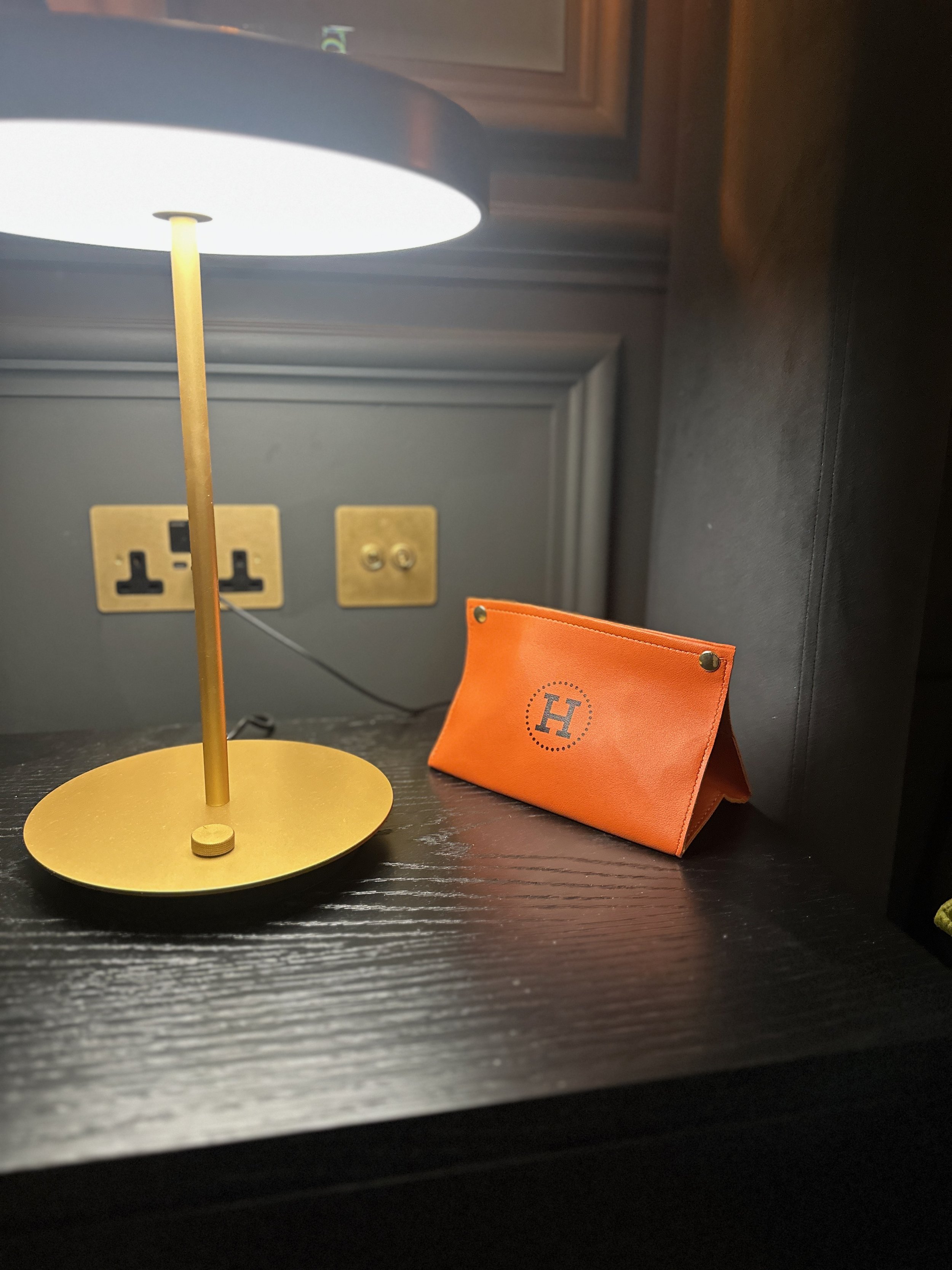 A black bedside table with a gold desk lamp on the left, and a folded orange leather wallet with a gray letter 'H' on the right. There is a blue wall with electrical outlets in the background.