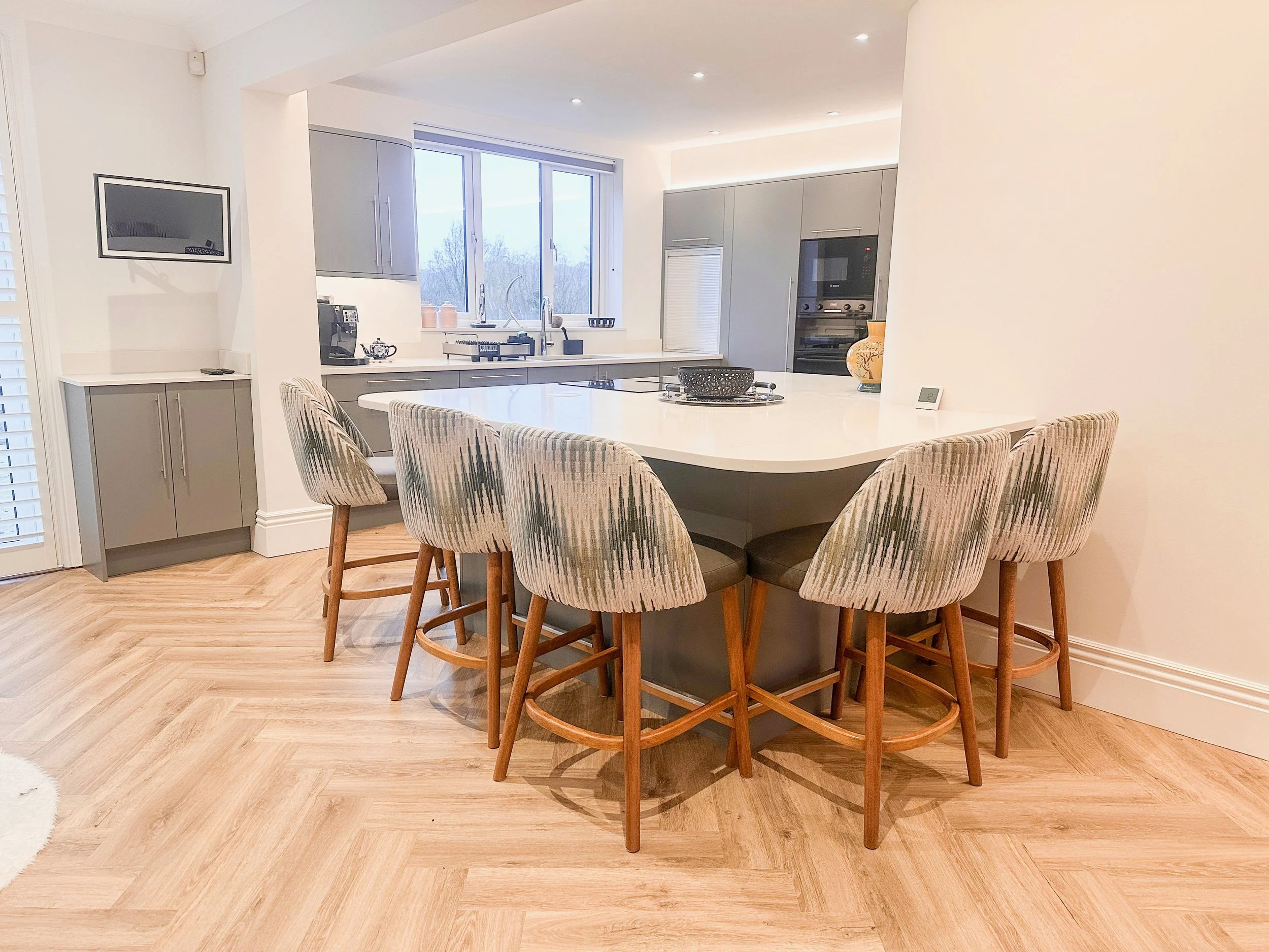 Modern kitchen with beige patterned chairs around a white island counter, gray cabinetry, and light wood flooring.