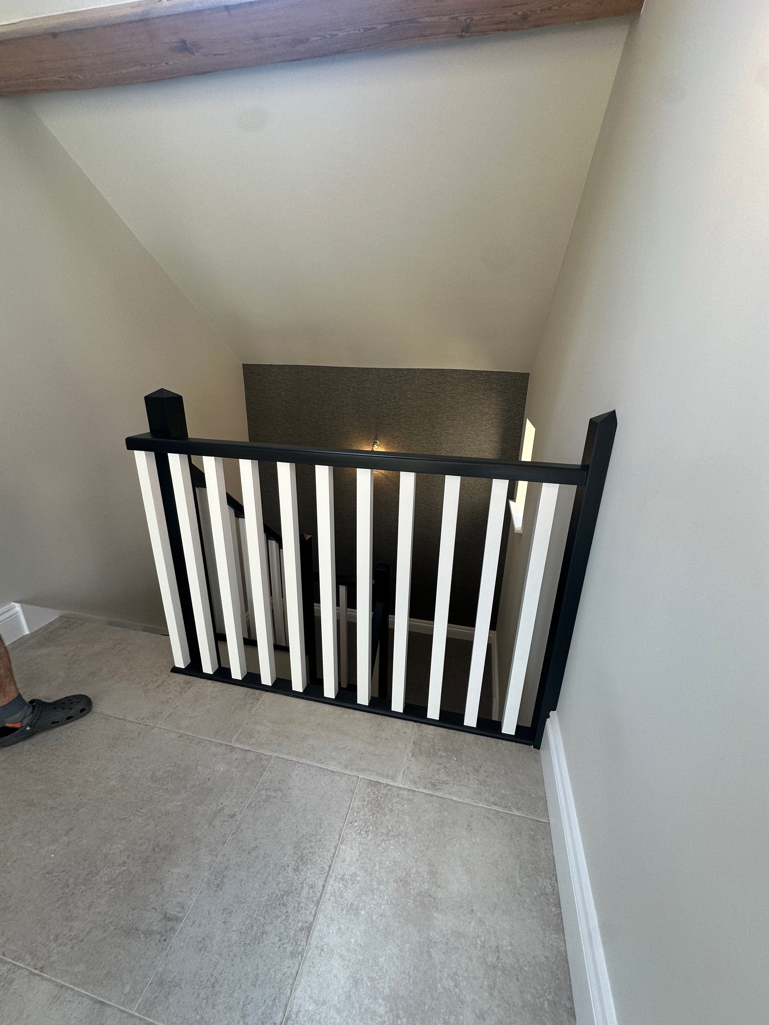 View of a staircase with a black and white wooden railing, leading down to a lower level with a textured dark wall and a wall sconce light. Part of a person wearing grey Crocs is visible on the left.