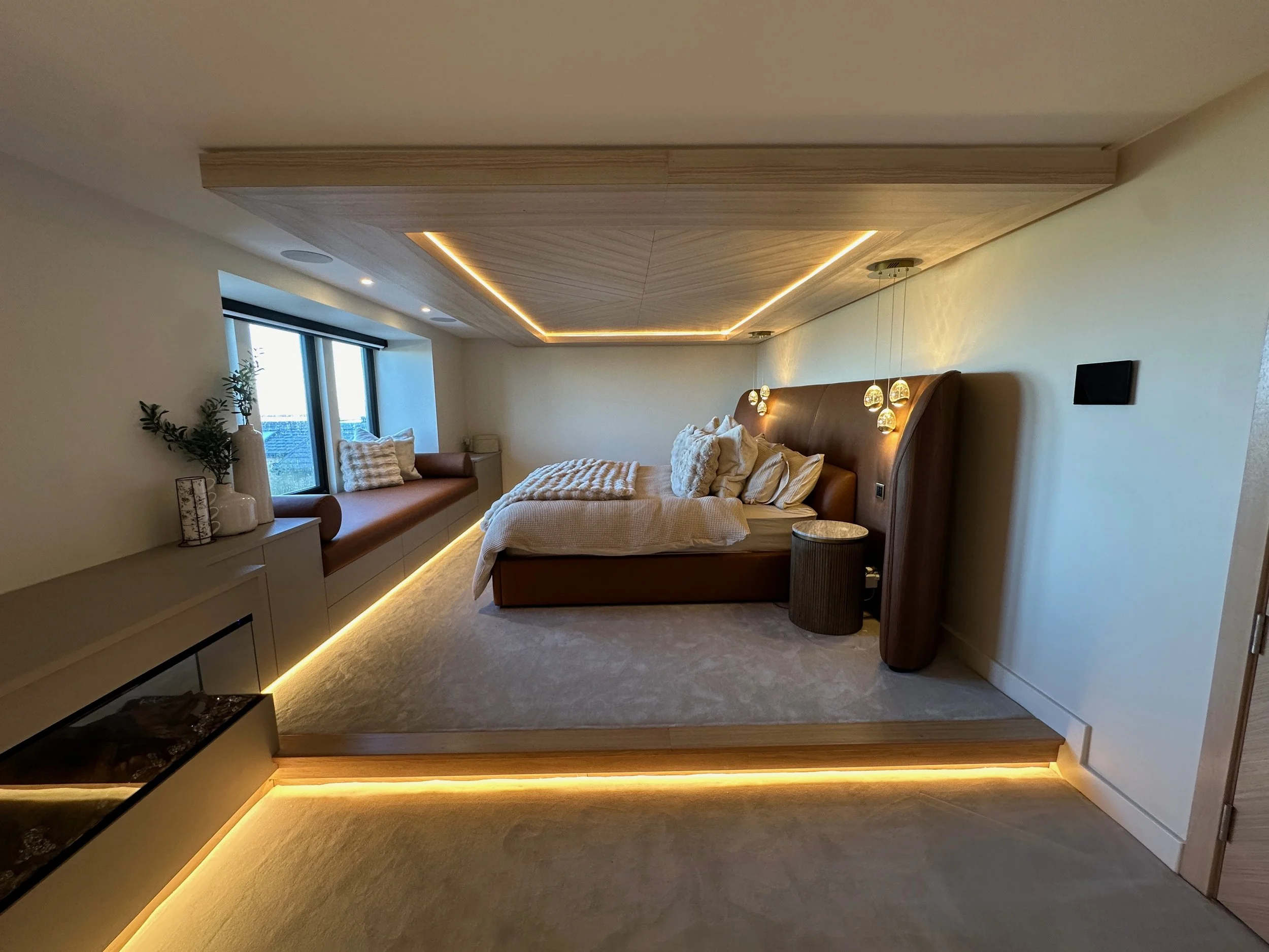 Modern bedroom with a large bed, decorative pillows, nightstand, pendant lights, window seat with pillows, and natural lighting.