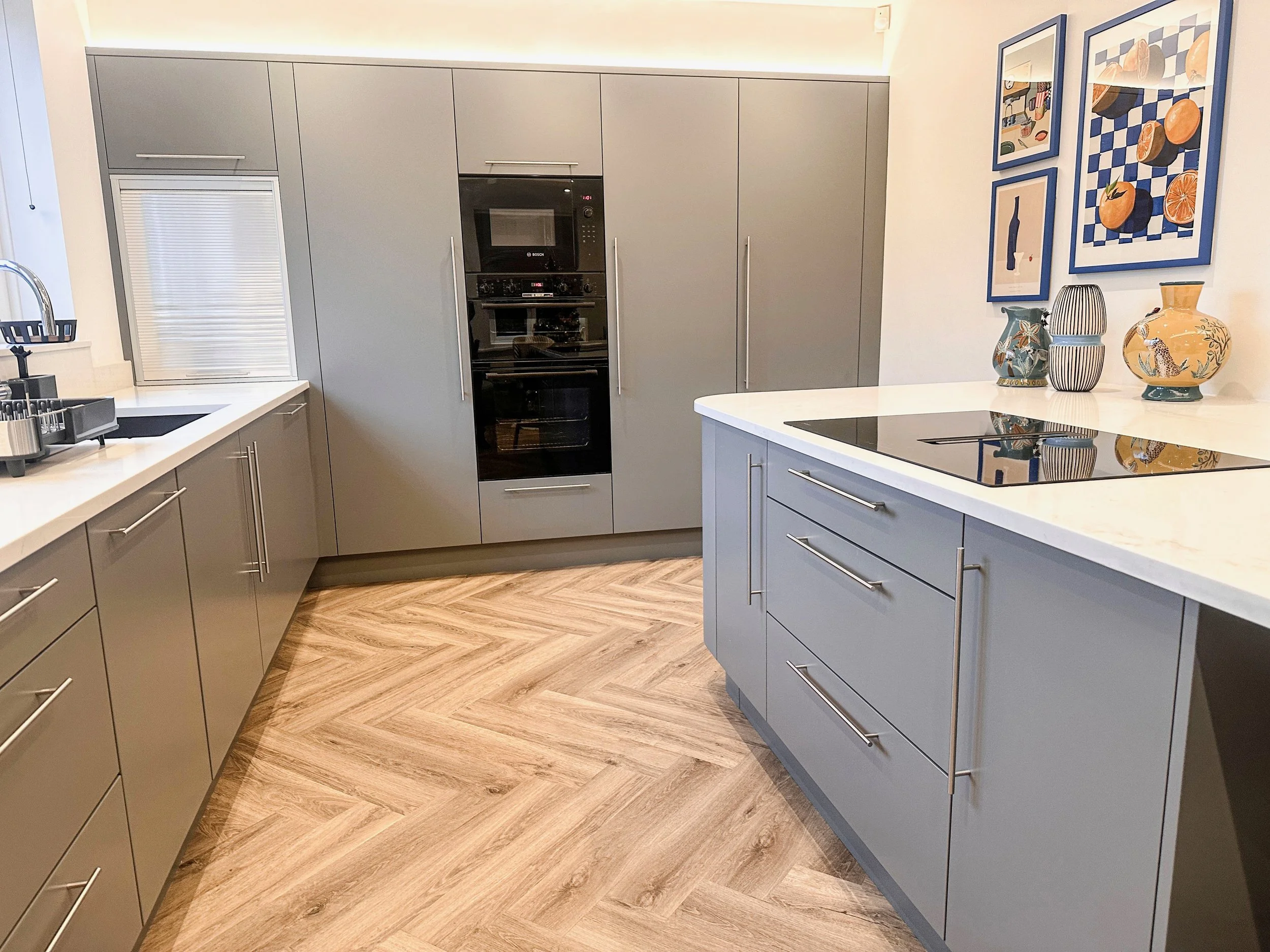 Modern kitchen with gray cabinets, white countertops, and wooden herringbone floor. Features a built-in oven, stovetop, and decorated vases on the counter. Wall art with orange-themed images hangs above.