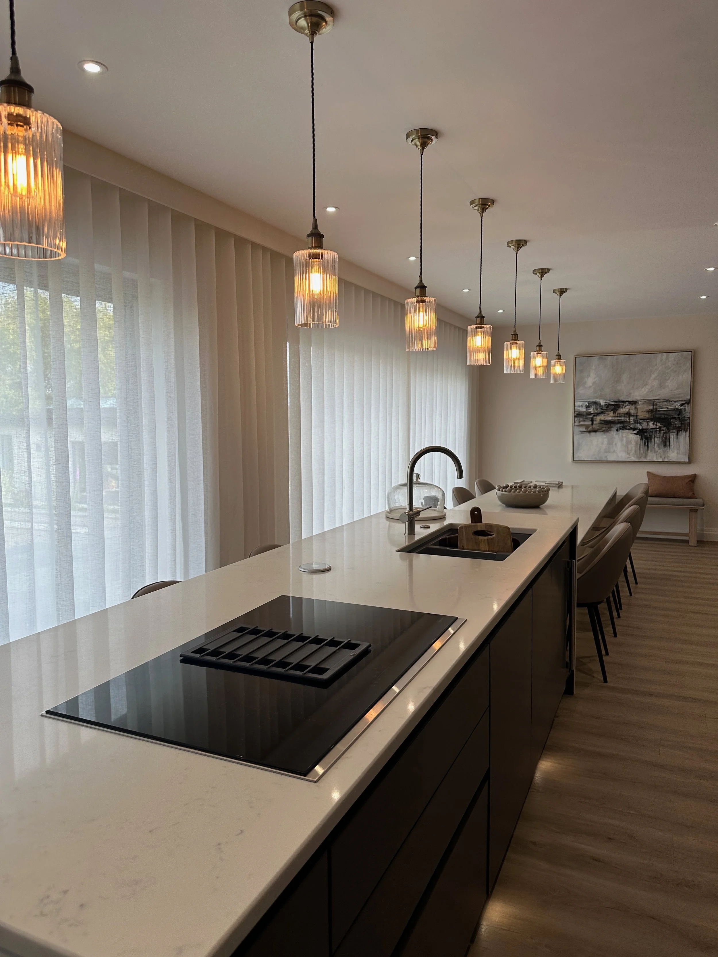Modern kitchen with a long white island counter, black stovetop, and a sink with a curved faucet. Overhead pendant lights hang in a row, and large windows with sheer curtains let in natural light. There is a piece of abstract art on the wall and a bench with a pillow in the background.