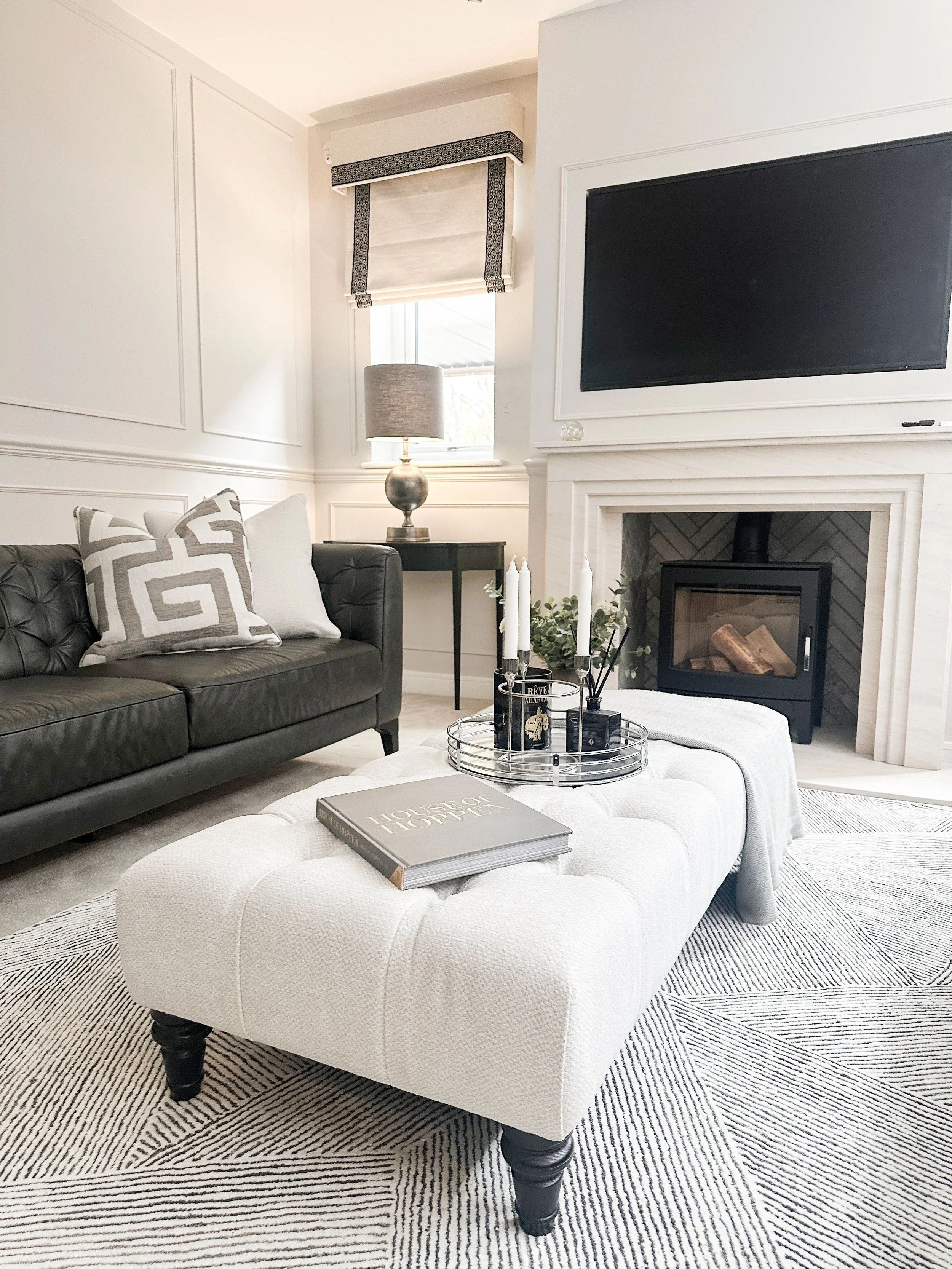 Living room with a black leather sofa, a white tufted ottoman with a book and tray on top, a fireplace with a black insert, a wall-mounted TV above the fireplace, a table lamp on a small black side table, and a window with a Roman shade.