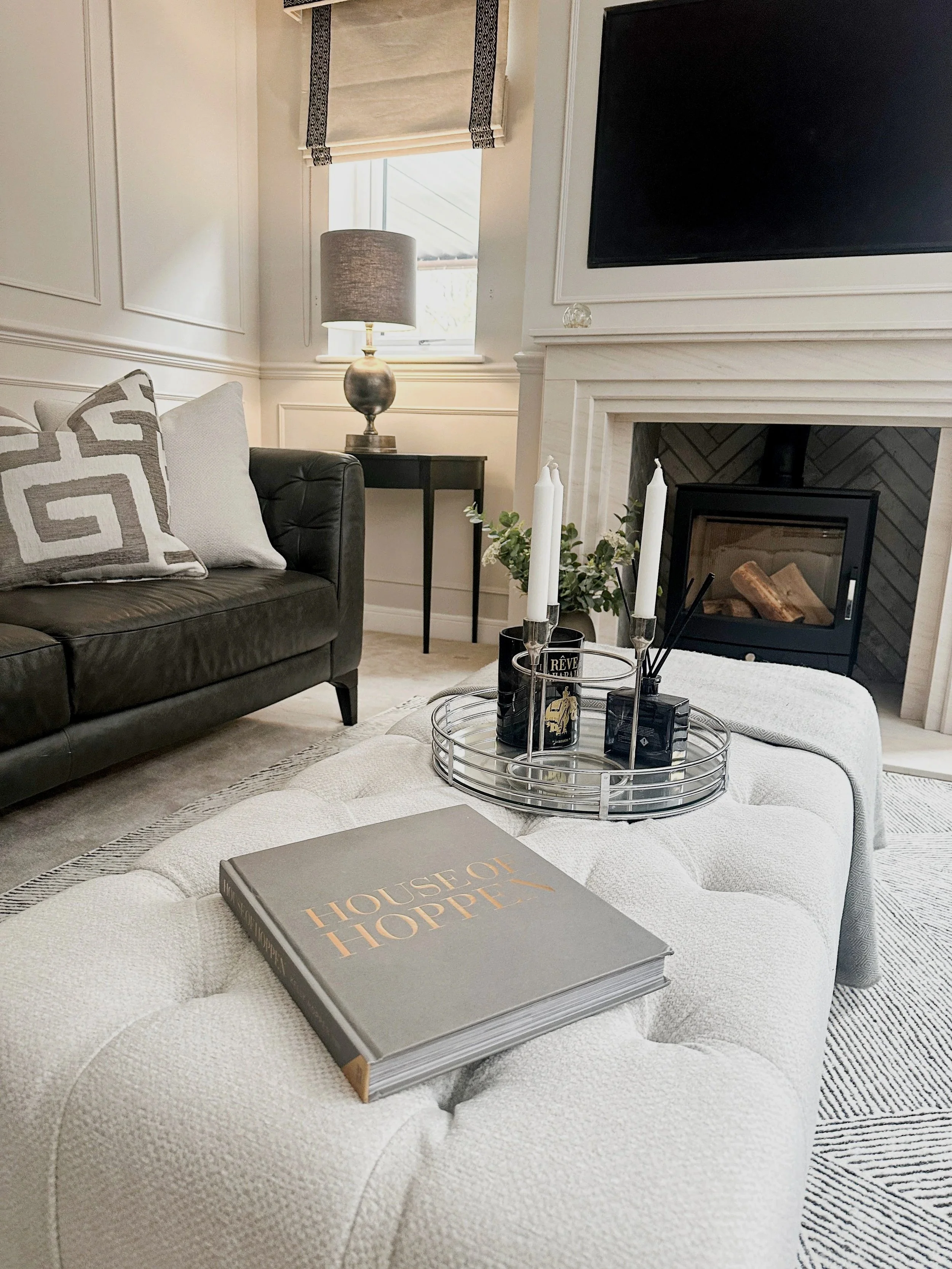 A cozy living room with a white upholstered ottoman, a black leather sofa with patterned pillows, a fireplace with logs, a flat-screen TV mounted above the mantle, a side table with a lamp, and decorative items on the ottoman and around the room.