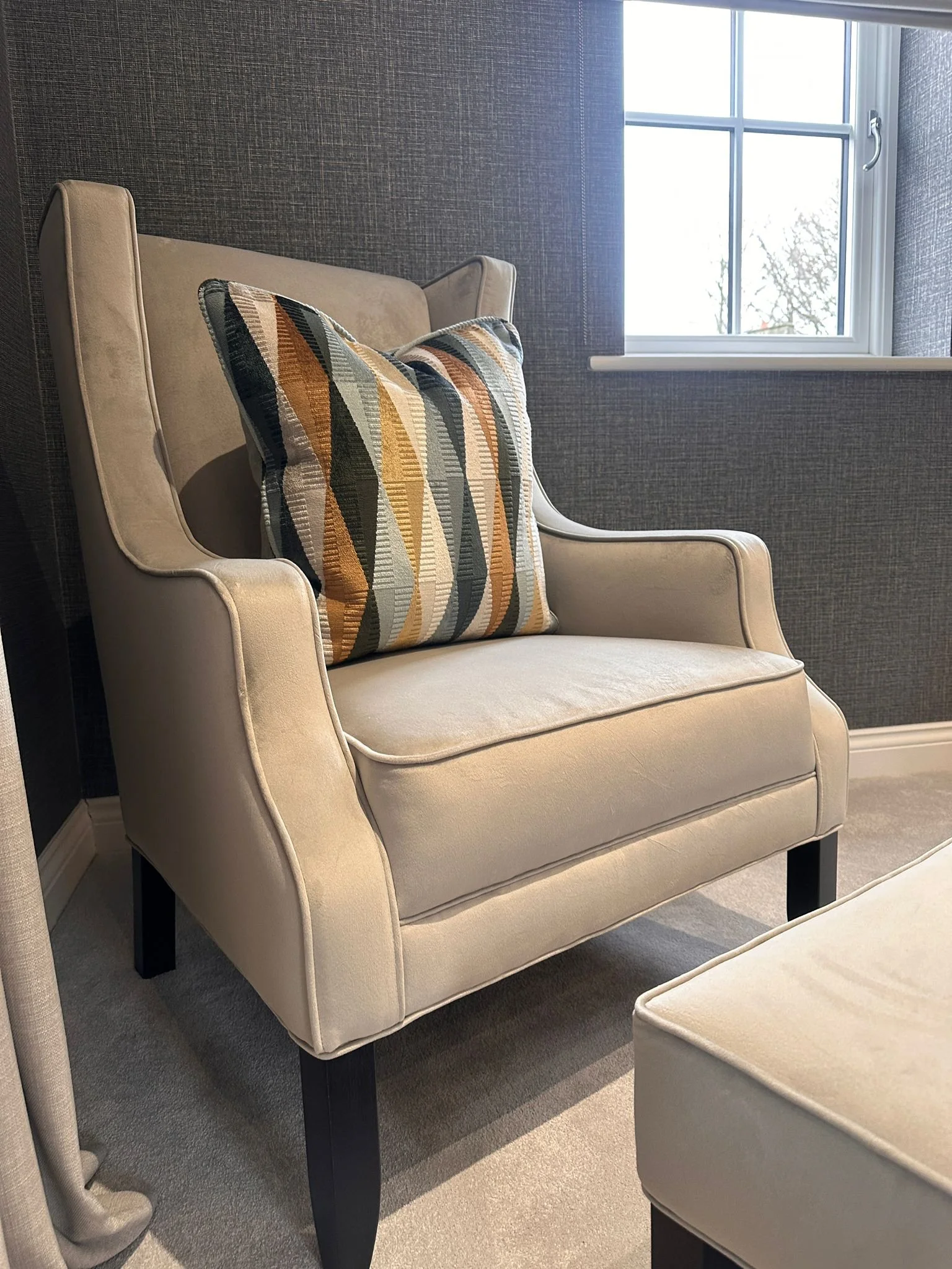 A beige armchair with a geometric-patterned pillow, placed near a window with a view of trees outside.