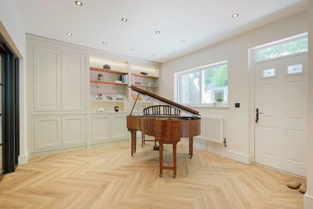 Empty room with a grand piano, built-in white cabinets with shelves, large window, white door, and wooden floor.