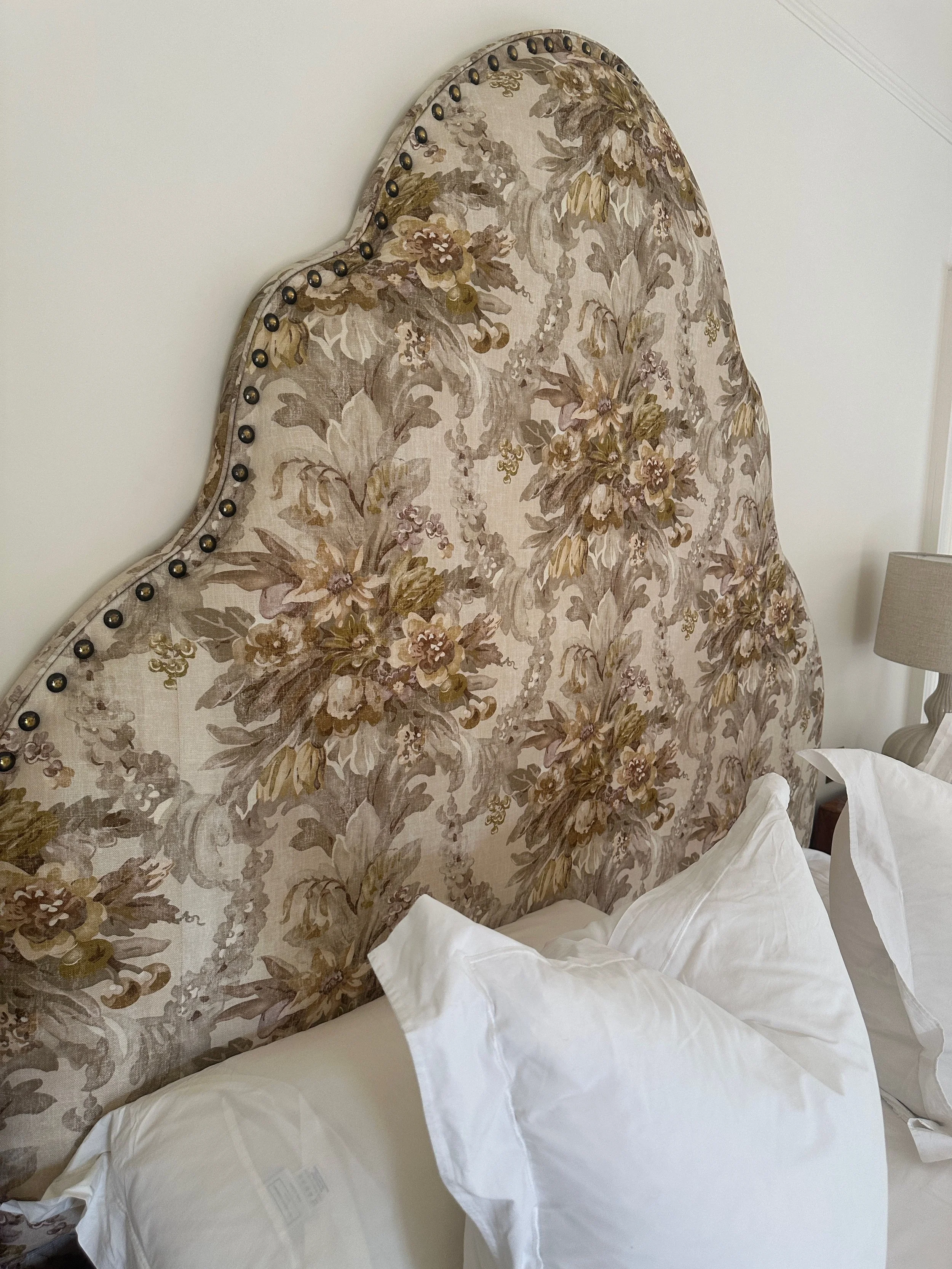 A floral upholstered headboard with beige, green, and pink tones, embellished with a row of brass nailhead trim, positioned behind a bed with white pillows and linens, next to a bedside table with a beige lampshade.