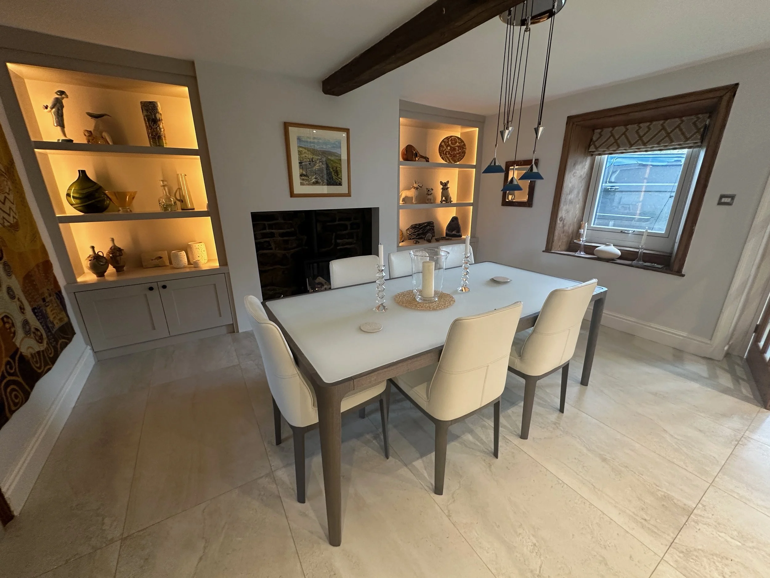  dining room with a white table and six cream-colored chairs, with a candle centerpiece, in front of a brick fireplace, flanked by two shelves with decorative items, a window with a brown wood frame, and a chandelier hanging above the table.