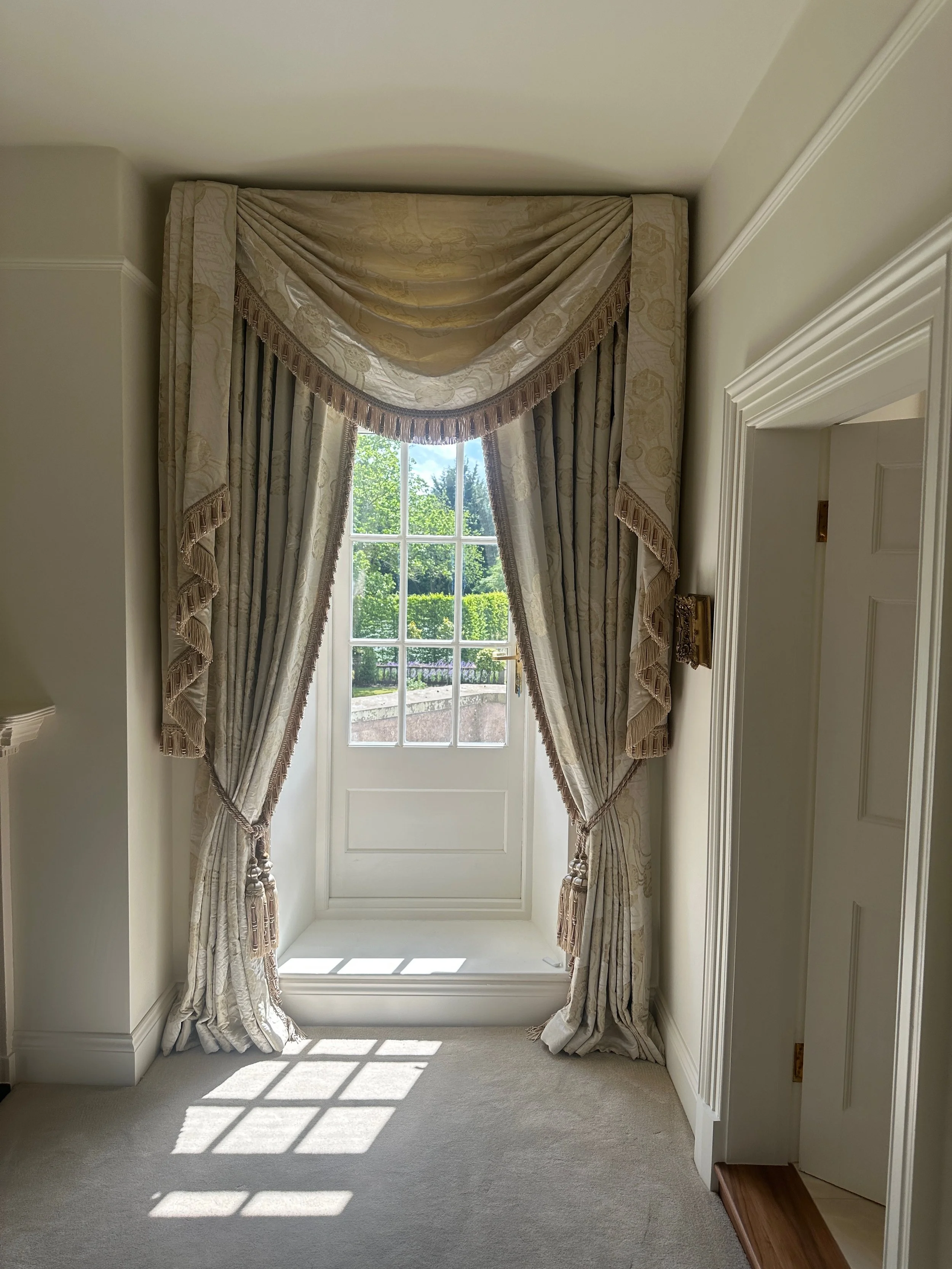 Elegant cream-colored curtains with tassels framing a glass door with a view of greenery outside, sunlight casting a pattern on the carpet.