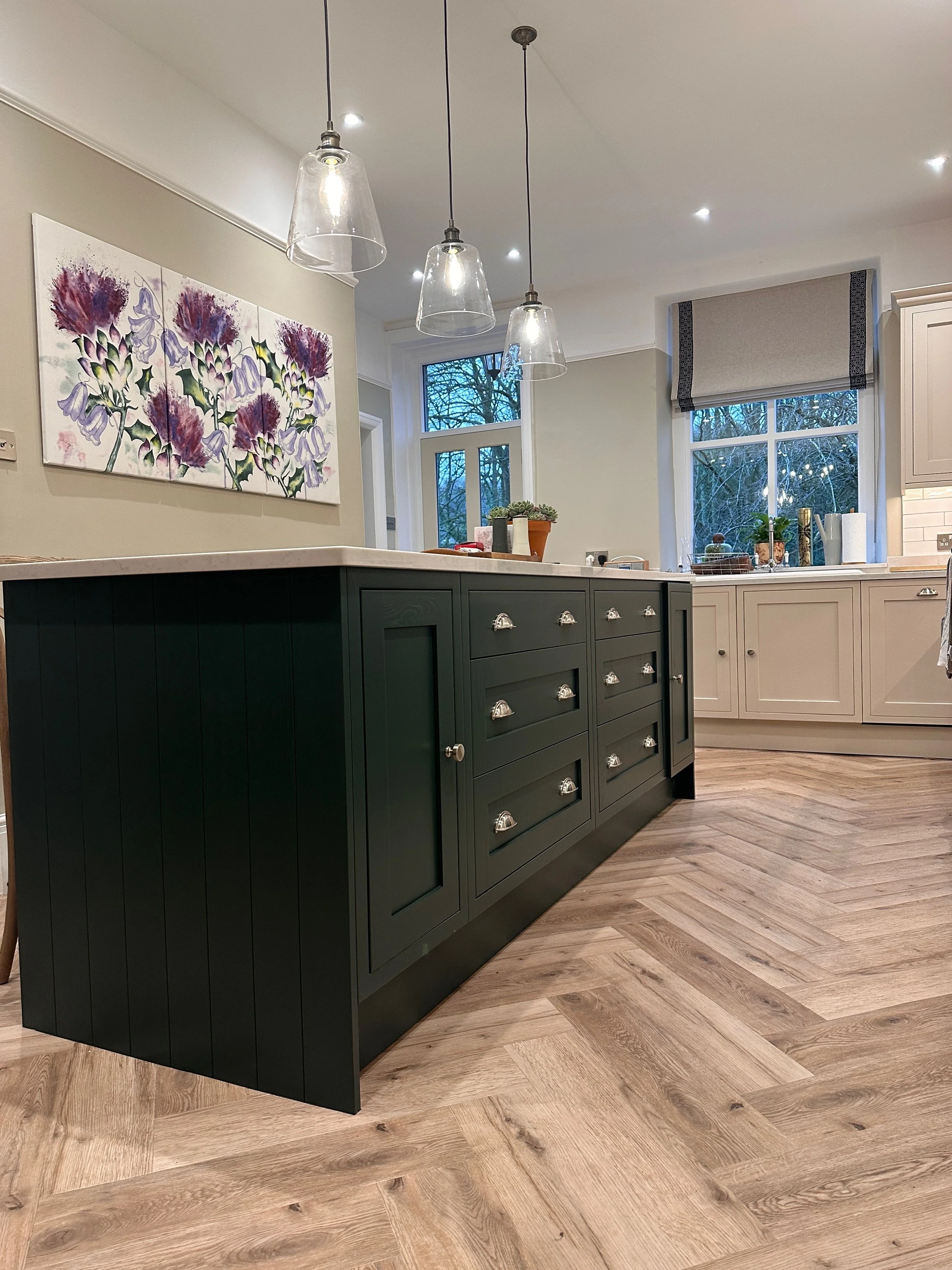 Modern kitchen with a white marble countertop island, dark green cabinets with silver handles, and light wood herringbone flooring. There are three glass pendant lights hanging above the island. Part of a floral painting hangs on the wall, and window