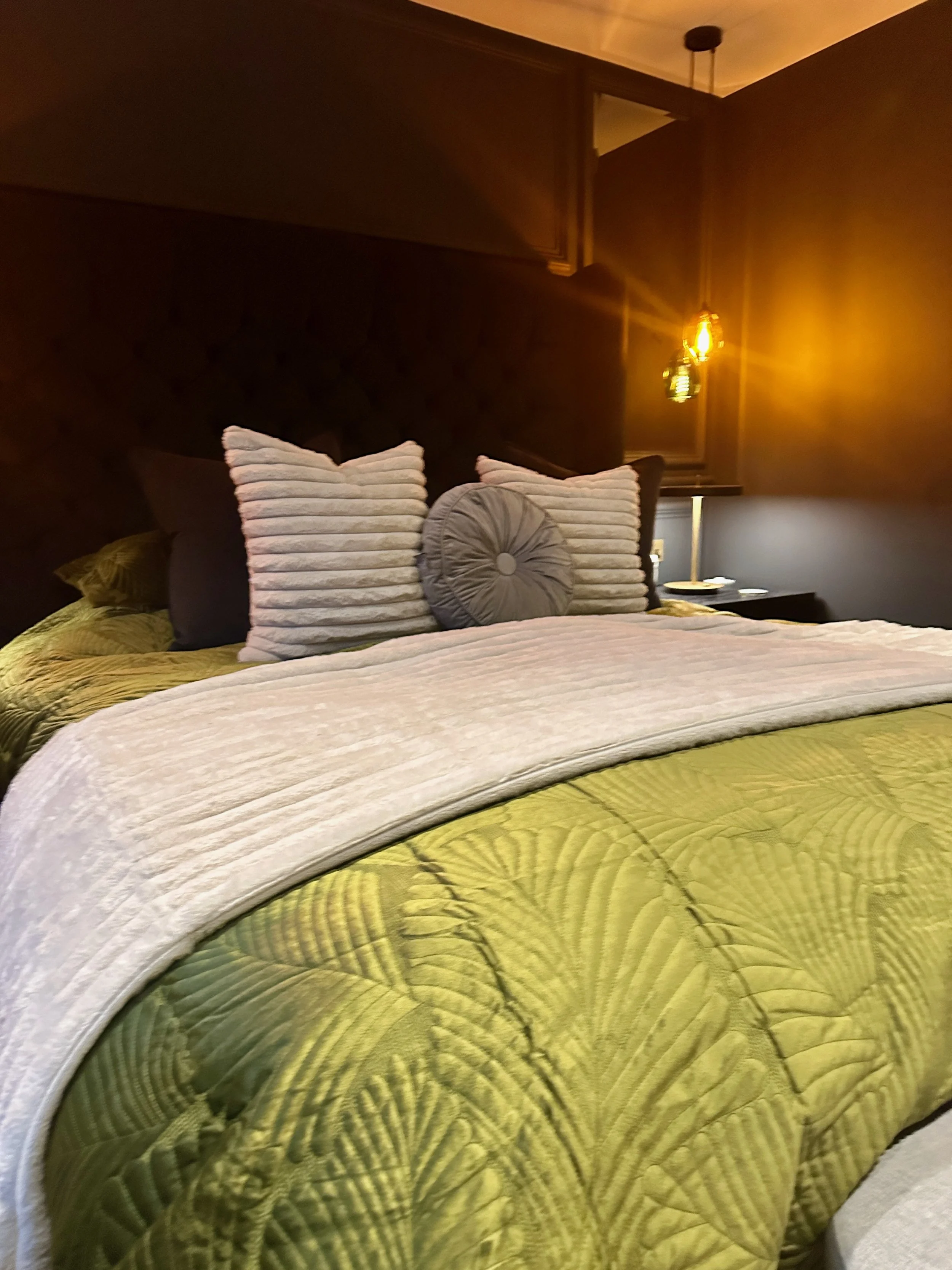 A neatly made bed with green tropical leaf-patterned bedding, topped with a folded white blanket and three decorative pillows in front, two beige striped and one round gray. A dark wooden headboard and side table with a modern lamp and some items are