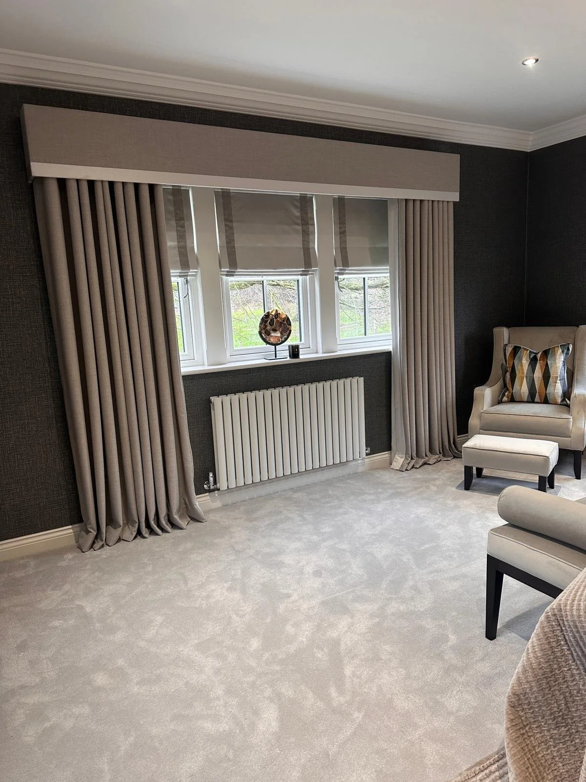 Living room with three windows, beige curtains, a beige armchair with a pillow, and a small beige ottoman.