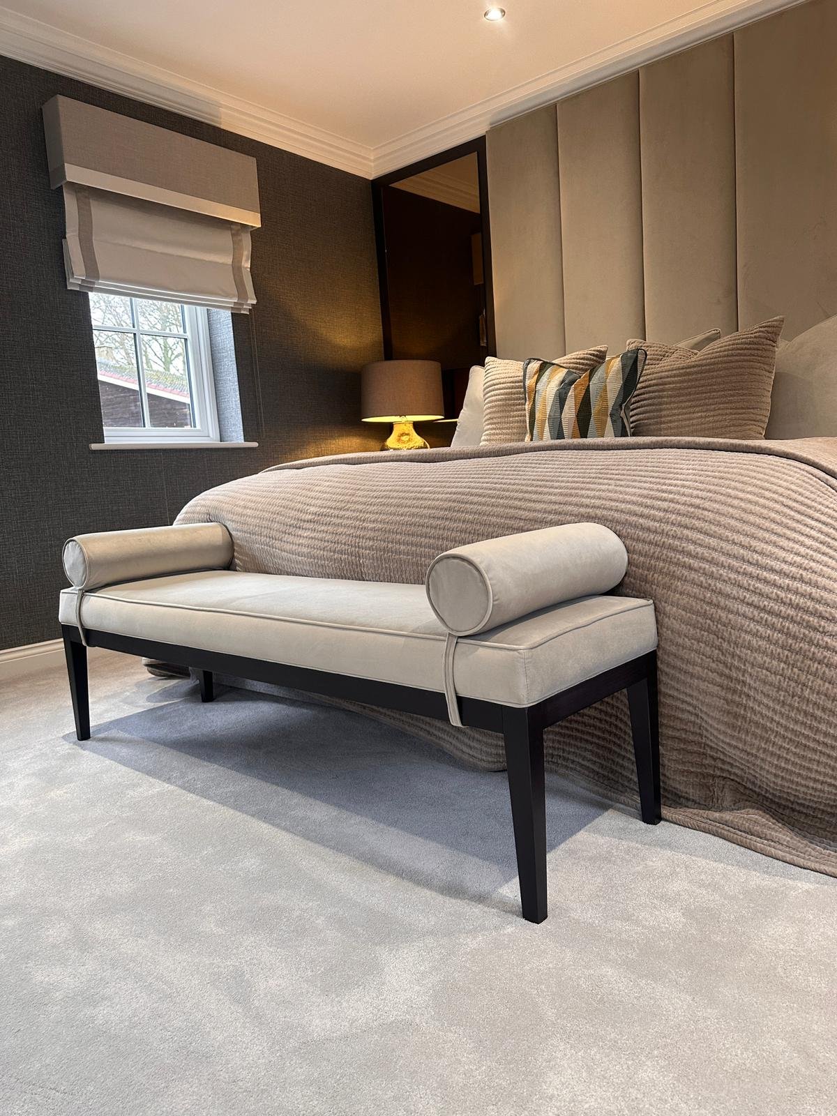 A padded bench with black legs and cylindrical pillows at each end sits in front of a bed in a bedroom.